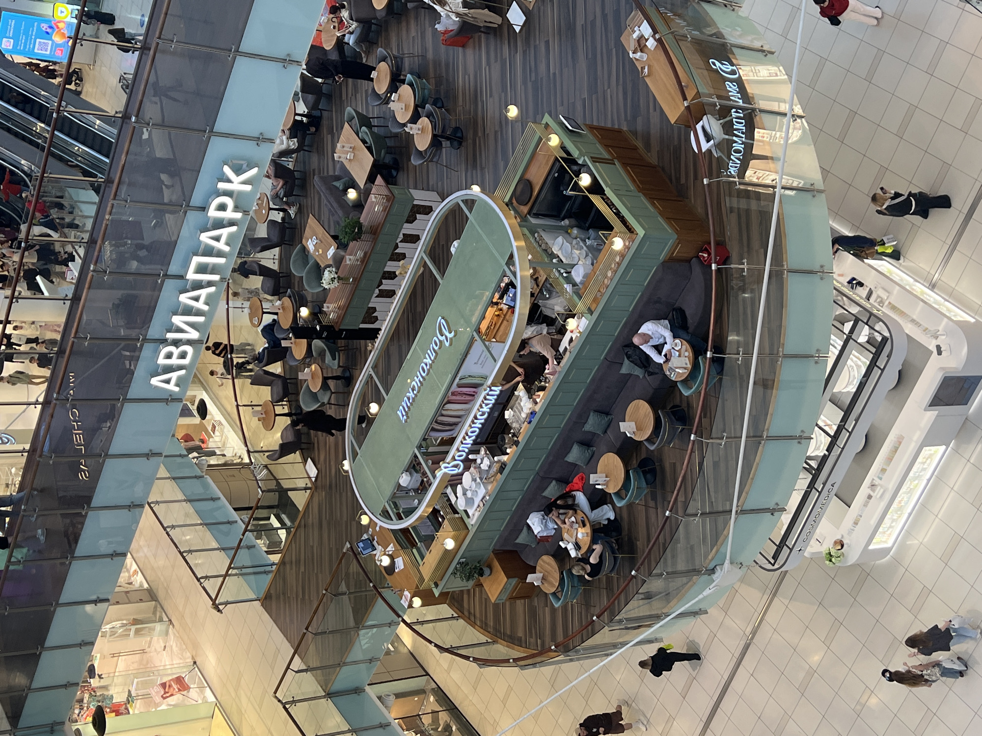 This is a high-angle, full-shot image of the interior of a modern, multi-level shopping mall. The mall is branded with the name "Aviapark," indicated by large white lettering on a blue background visible across the upper level. The architecture features glass railings, wooden floors, and escalators.
In the mid-ground, there's a circular, green-colored kiosk or cafe with seating areas. The kiosk has a counter and displays. Two individuals are seated at a small table in front of this establishment. The name "Волконский" (Volkonsky) is prominently displayed on the green structure.
On the upper level, there are several cafes and seating areas with people sitting at tables and on escalators. The scene suggests a busy commercial environment.
In the foreground and lower levels, there are various shops with displays and more people walking around. One shop in the lower foreground has a white counter with illuminated displays and the name "COSMOMEDICA" visible.
The overall impression is that of a bustling shopping center, possibly during daytime hours, with a diverse range of commercial activities and shoppers. The location is identified as Aviapark, Khimki, Russia.