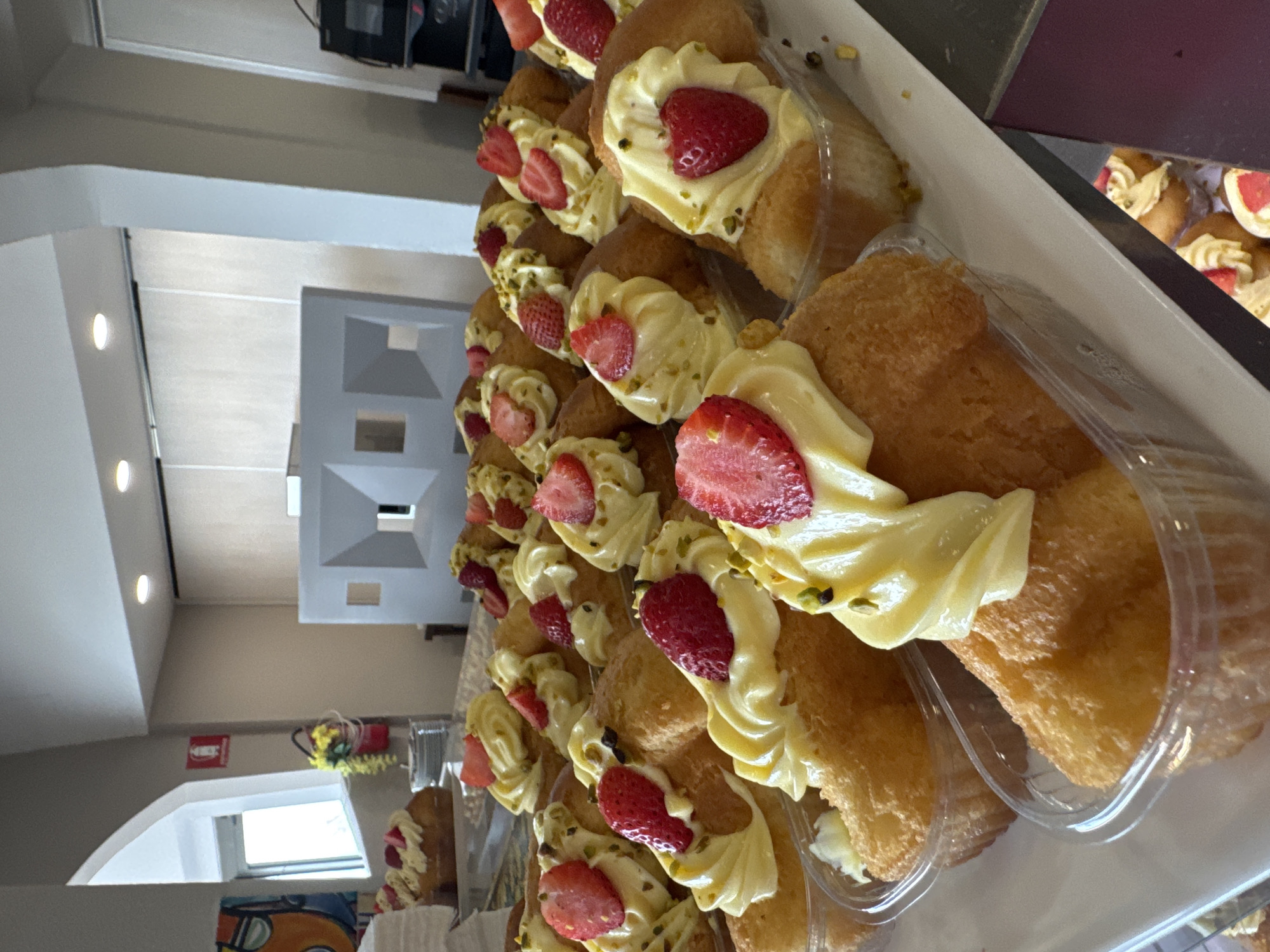 The image depicts a display of individually packaged pastry desserts, likely in a bakery or confectionery shop located in Isola delle Femmine, Italy. The pastries are small, golden-brown, cake-like bases topped with a swirl of pale yellow cream, a slice of fresh strawberry, and a sprinkle of chopped green nuts, possibly pistachios. They are arranged in clear plastic containers on a white tray. The background shows a brightly lit interior with a modern, angular white partition or display unit, soft lighting from overhead, and a window with an arched top to the left, revealing an outdoor scene. A red fire extinguisher is visible on the wall near the window. There are no people visible in the frame.