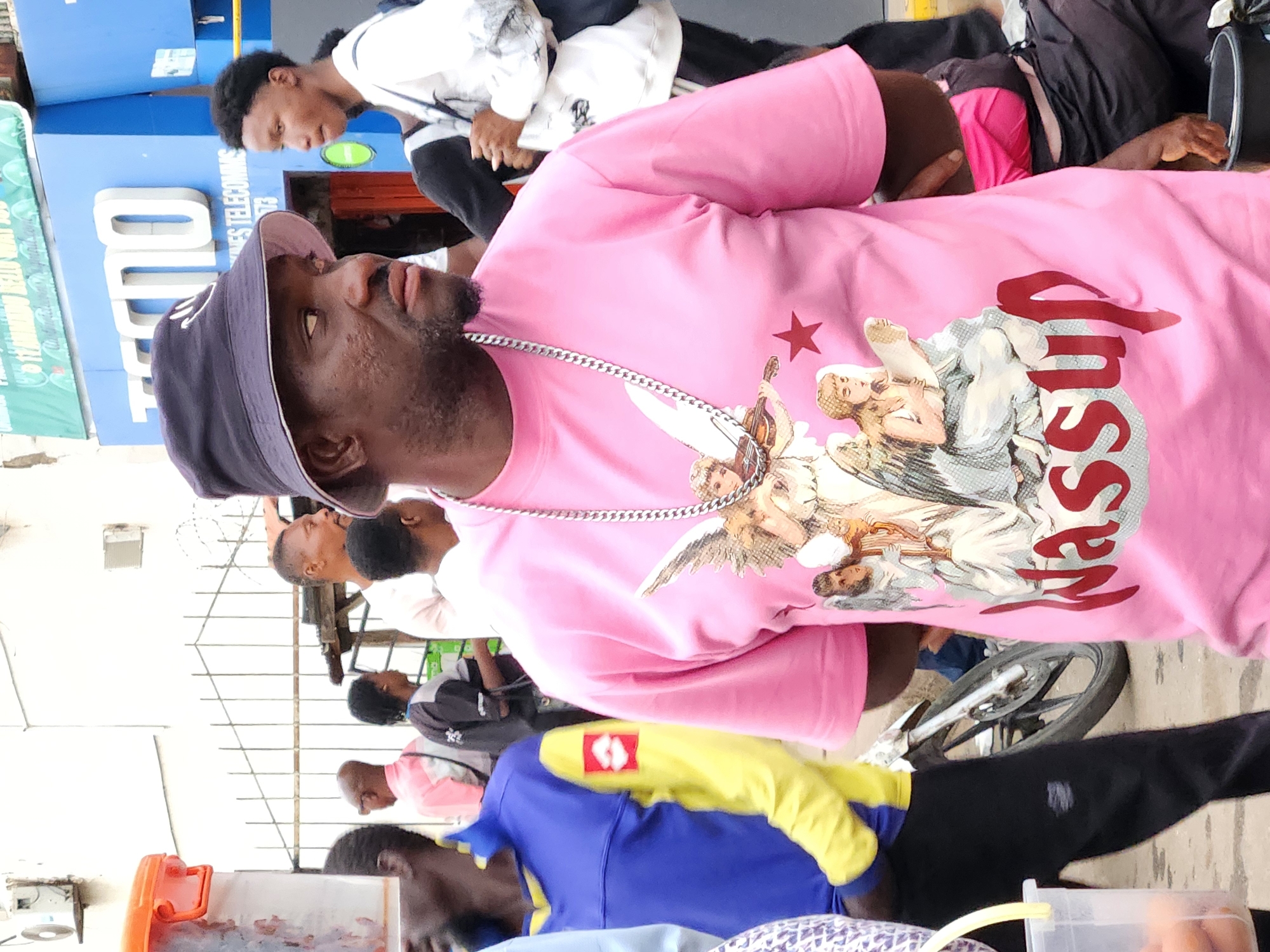 The image shows a group of people in what appears to be an urban environment, likely in Jos, Nigeria. The central figure is a man wearing a pink T-shirt with an angel graphic and the word "Senasup" on it, along with a black bucket hat and a silver chain. He is surrounded by other individuals, some wearing casual clothing. The background includes a building with the word "DIO" on it. A bicycle and various containers, possibly containing food or drinks, are also visible in the scene.
