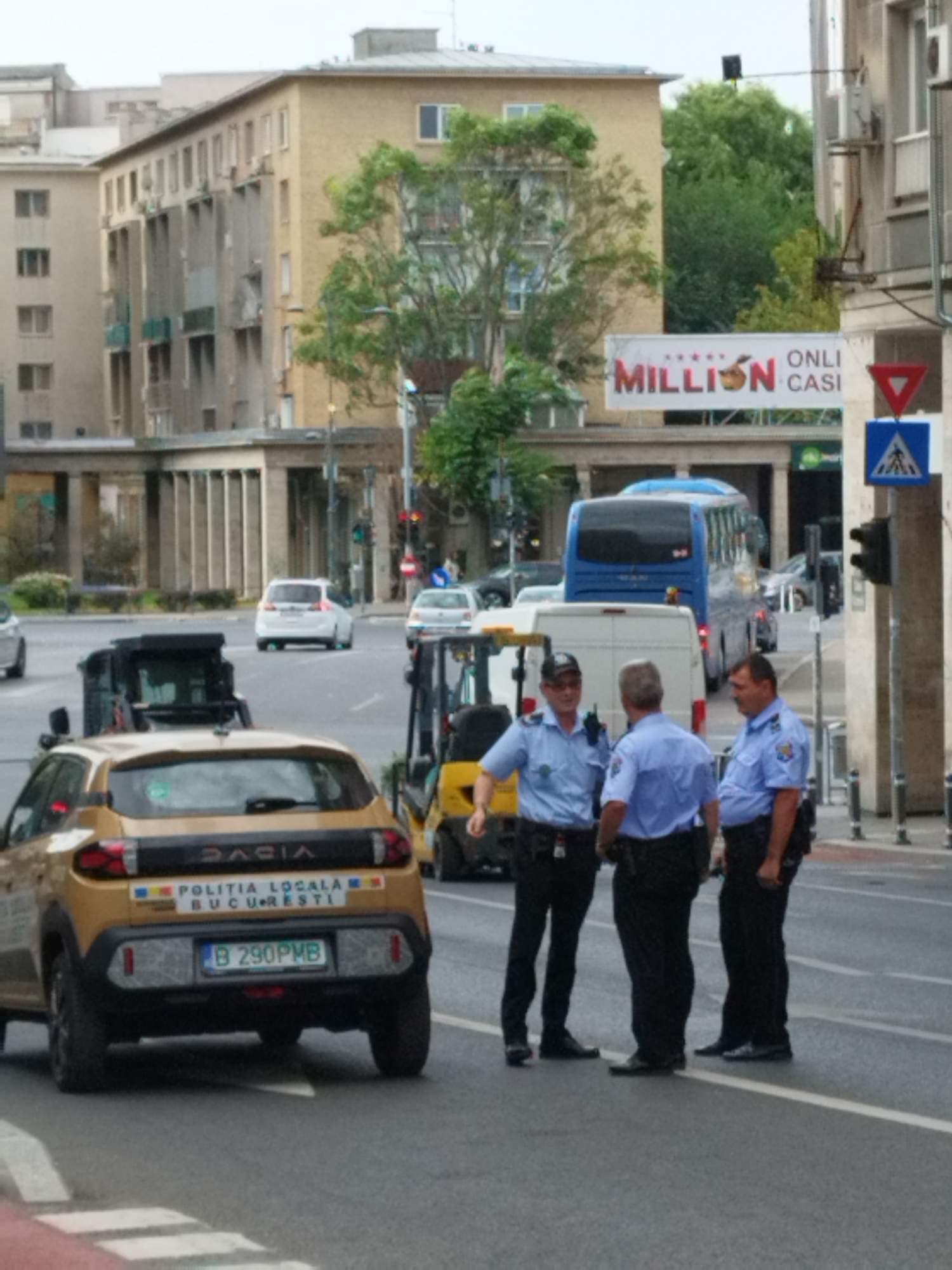 The image depicts a street scene in Bucharest, Romania, featuring several individuals in police uniforms, vehicles, and urban infrastructure. In the foreground, a gold-colored Dacia vehicle, identified by license plate "B 290PMB" and "POLITIA LOCALA BUCURESTI" markings, is parked on the street. Three uniformed officers are present near a white van and a yellow forklift. The officer on the left, wearing a cap and a blue shirt, appears to be speaking. The middle officer, also in a blue shirt, has his back turned. The officer on the right, in a blue shirt, is facing away from the camera.
In the background, a blue bus is visible, along with other vehicles and buildings. A large billboard with "MILLION CASI" is displayed on one of the buildings. Traffic signals and pedestrian crossing signs are also present, indicating an intersection. The environment is an urban street with paved roads and sidewalks. The overall atmosphere suggests an ongoing police presence or operation.