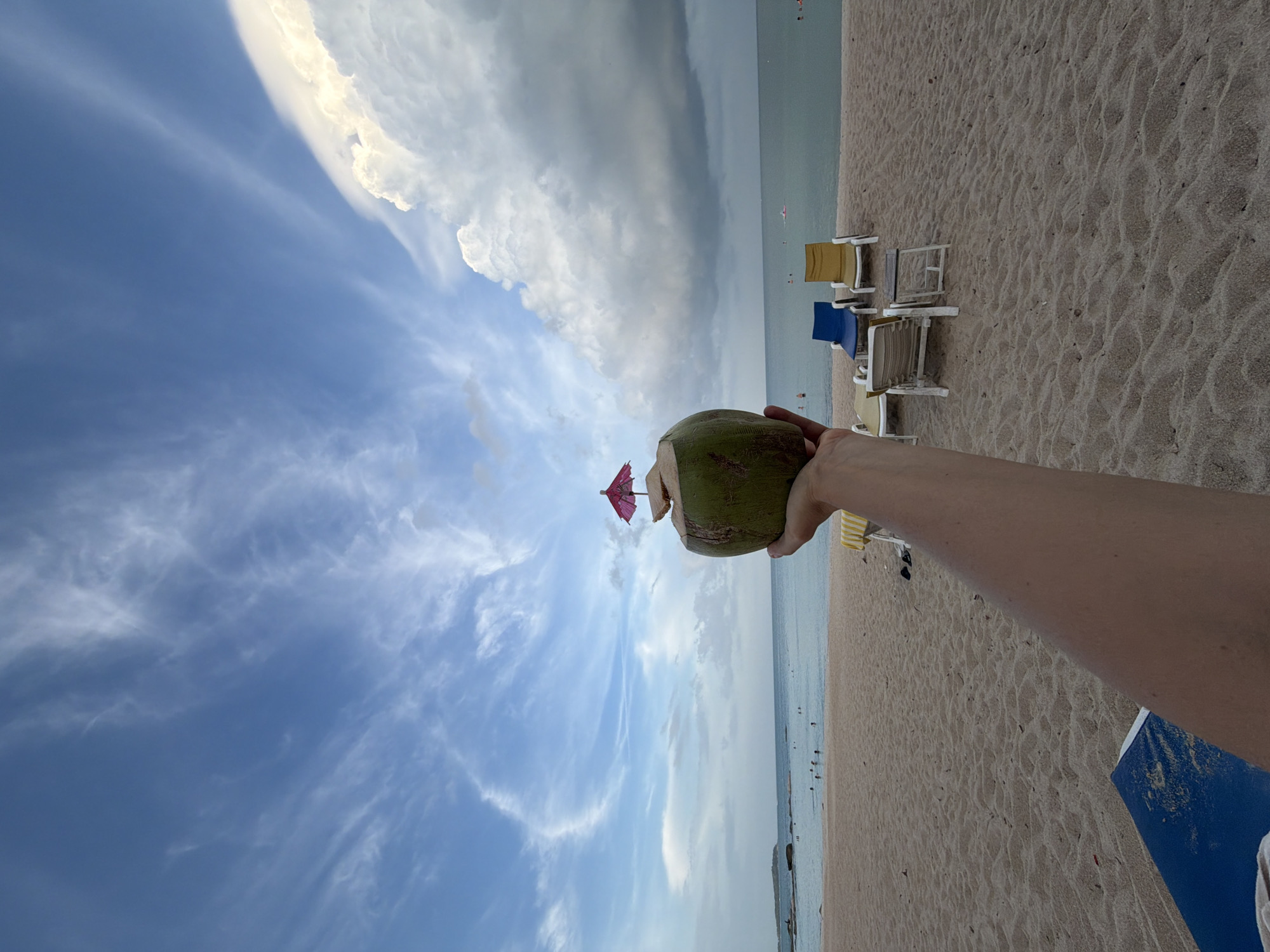 A person's arm extends into the frame, holding a young coconut with a small pink umbrella inserted into it, positioned in the foreground. The coconut is the primary subject. Behind it, a sandy beach stretches towards the ocean. Several lounge chairs, some blue and yellow, others white, are scattered on the sand. In the distance, people are visible in the calm, turquoise water, swimming and relaxing. The horizon separates the sea from a sky filled with dramatic, wispy white clouds against a blue backdrop. The overall scene depicts a tropical beach setting, likely on the island of Ko Samui, Thailand, known for its picturesque beaches and clear waters.