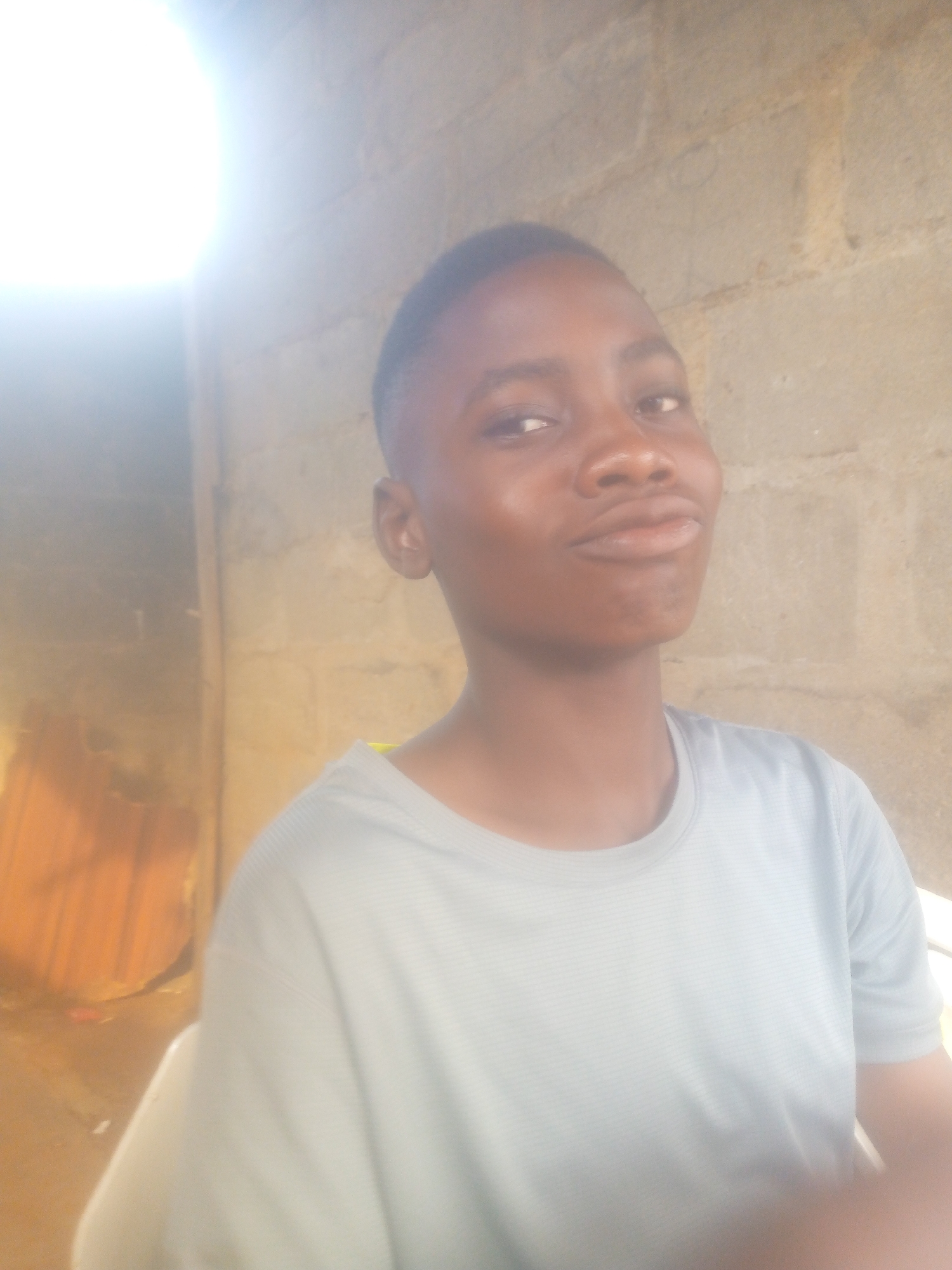 A waist-up portrait features a young person with dark skin, short dark hair, and brown eyes, looking directly at the camera with a subtle, slightly playful or smirking expression. The individual is wearing a light blue-grey, short-sleeved t-shirt with faint horizontal striping.
A strong, overexposed light source, likely natural sunlight, is visible in the upper left corner, casting a bright glare and illuminating the subject from that side. The background to the right of the subject consists of a rough, light brown or beige wall, appearing to be constructed from cinder blocks or similar masonry. On the left side of the background, beneath the bright light, there's a darker, warmer-toned section, possibly a wooden or metallic surface, or another part of the building.
The setting suggests an outdoor environment or an indoor space with ample natural light. The time of day appears to be daytime due to the bright sun. No text is visible in the image.