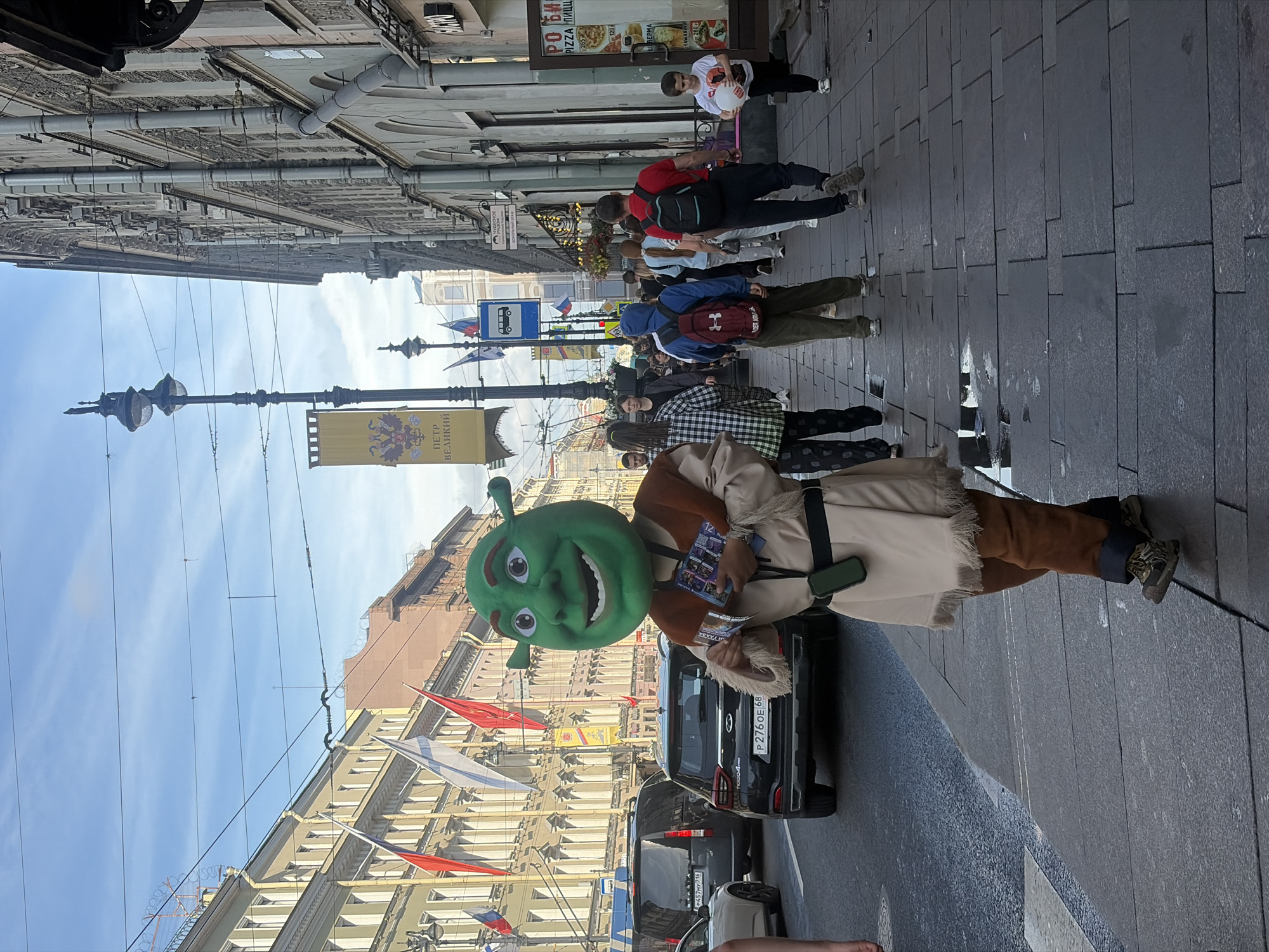 This media file captures a street scene in Saint Petersburg, Russia. The primary subject is a person in a Shrek costume, complete with a large green mask and brown and tan clothing. The Shrek character is standing on a sidewalk next to a black SUV. Behind them, other people are walking along the street, some carrying backpacks. The street is paved with large, dark gray tiles, and there are buildings lining both sides of the street, appearing to be shops or businesses.
Several flags are visible, including Russian flags and other decorative banners. One banner features a double-headed eagle emblem, often associated with Russian imperial symbolism, and the text "ПЕТР ВЕЛИКИЙ" (Peter the Great). Another banner above a shop has text that appears to be advertising pizza and shawarma. A blue bus stop sign is also visible in the background.
The scene suggests a daytime activity, likely an event or festival, given the presence of costumed characters and decorative banners. The sky is bright blue with some clouds, and the lighting indicates it is daytime, possibly afternoon. The ground appears slightly wet, suggesting recent rain or a cleaning. The overall atmosphere seems lively and public. The license plate on the black SUV is partially visible and reads "P276 OE 68".