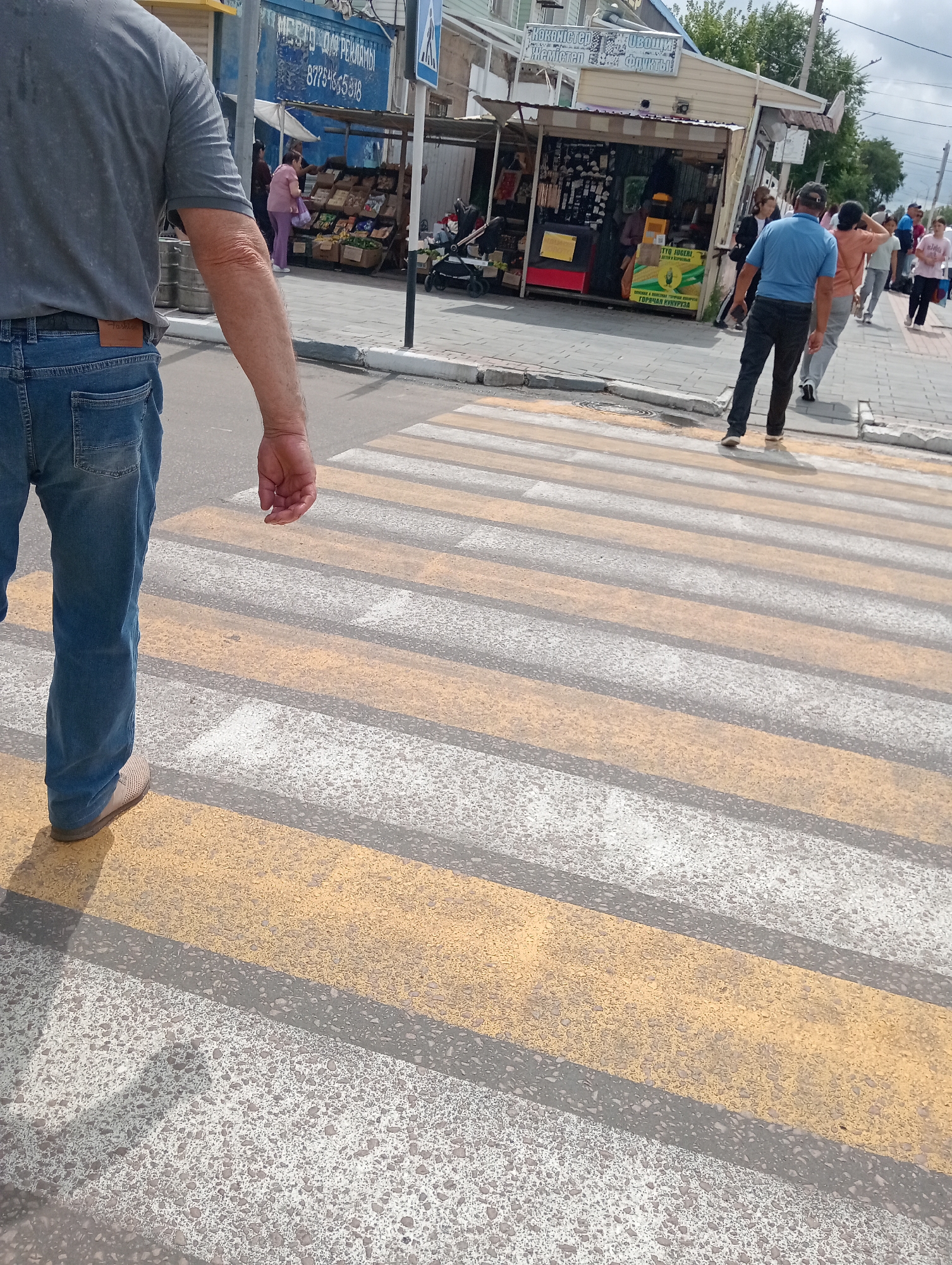 A man wearing a grey t-shirt and blue jeans walks across a crosswalk. The crosswalk is marked with alternating yellow and white stripes. In the background, a street market is visible with stalls selling goods. A few other people are walking on the sidewalk. The scene appears to be in a city in Kazakhstan.