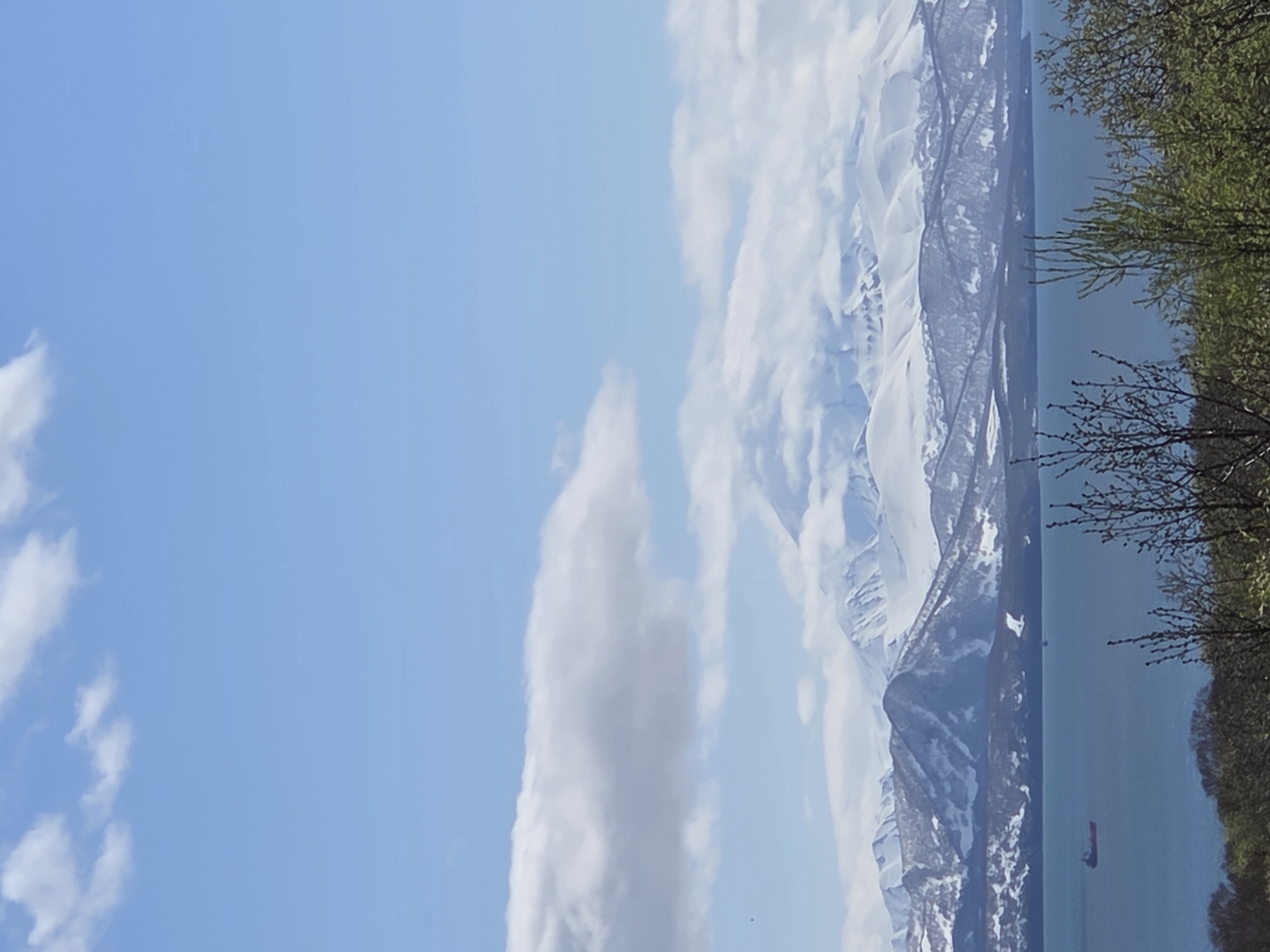 The image depicts a landscape under a blue sky with scattered clouds. A prominent, elongated cloud formation stretches vertically in the center. In the background, snow-capped mountains are visible, partially obscured by clouds. A body of water lies below the mountains, and foliage, possibly trees, lines the right edge of the image. A small boat is visible on the water. The location is Unknown, Unknown.