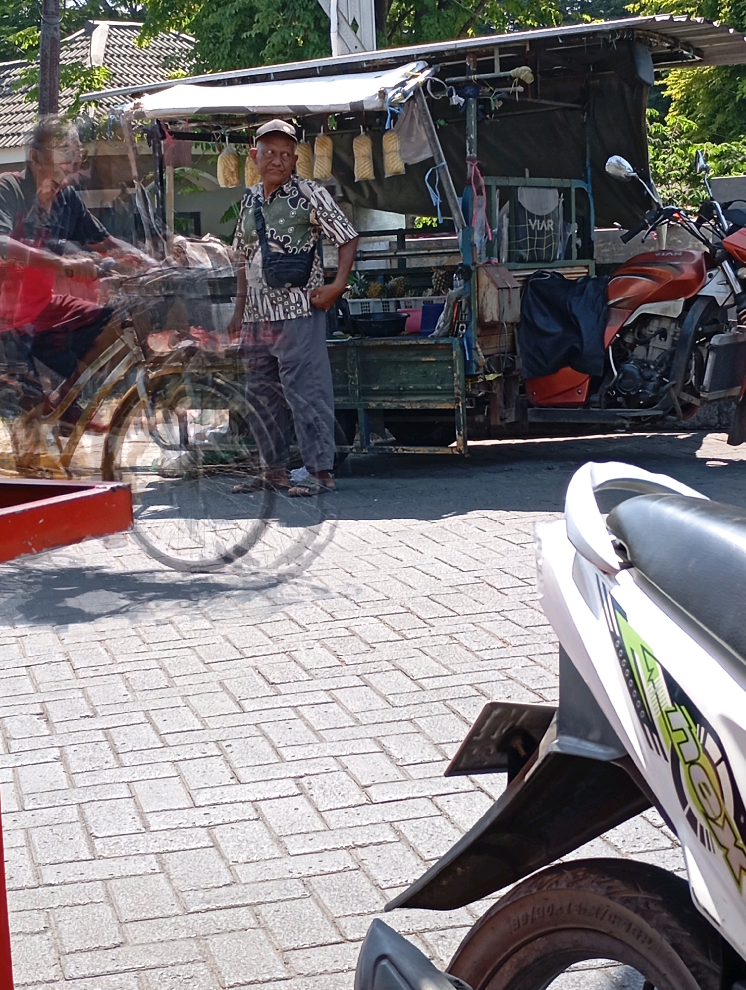 In Kalisampurno, Indonesia, an outdoor scene captures a street vendor and his cart. The vendor, a man wearing a patterned short-sleeved shirt, grey pants, and a cap, stands beside his merchandise cart which is adorned with various items. A partially blurred image of another person on a bicycle, dressed in a red and black shirt, is also visible in the foreground, suggesting movement. The cart itself appears to be a mobile setup with a covered roof, and a motorcycle is parked to the right, partially obscuring the cart. The ground is paved with interlocking bricks.