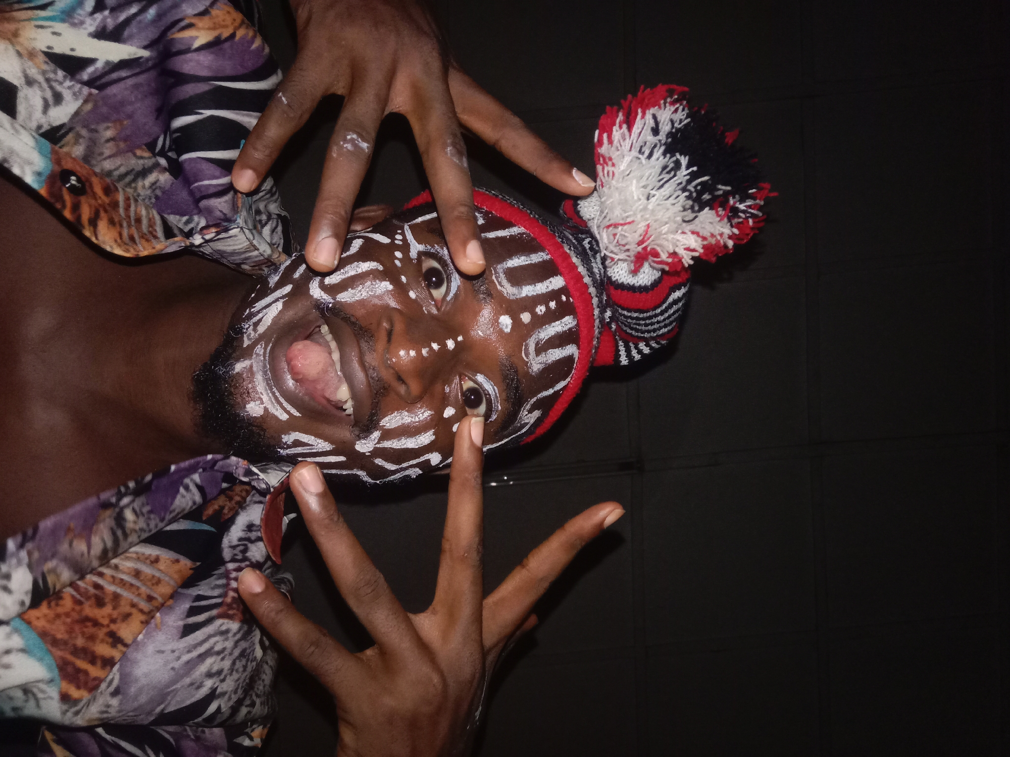 A vibrant, close-up portrait features a person with dark skin, their face adorned with striking white geometric patterns of dots and lines. The individual is captured in an expressive moment, looking directly at the viewer with wide eyes and an open mouth, suggesting they are shouting, laughing, or making a strong vocalization. They wear a red, black, and white knitted cap, featuring a striped pattern and a large, fluffy pompom of matching colors on the top right side of their head.
Two dark-skinned hands frame the person's face, with fingers spread and some white paint residue visible on the fingertips, indicating interaction with the face paint or a gestural expression. The person is also wearing a colorful, patterned garment or shirt with abstract shapes and colors including purple, brown, black, and white, visible on their shoulders and upper chest.
The background is uniformly dark, creating a stark contrast that isolates and highlights the subject. Subtle lines in the dark background might suggest a tiled or paneled wall. The image is oriented dynamically, with the person's head tilted to the left, contributing to the energetic and immediate feel of the photograph. The overall scene conveys a sense of celebration, performance, or cultural display, characterized by strong emotion and distinctive attire. No text is visible in the image.