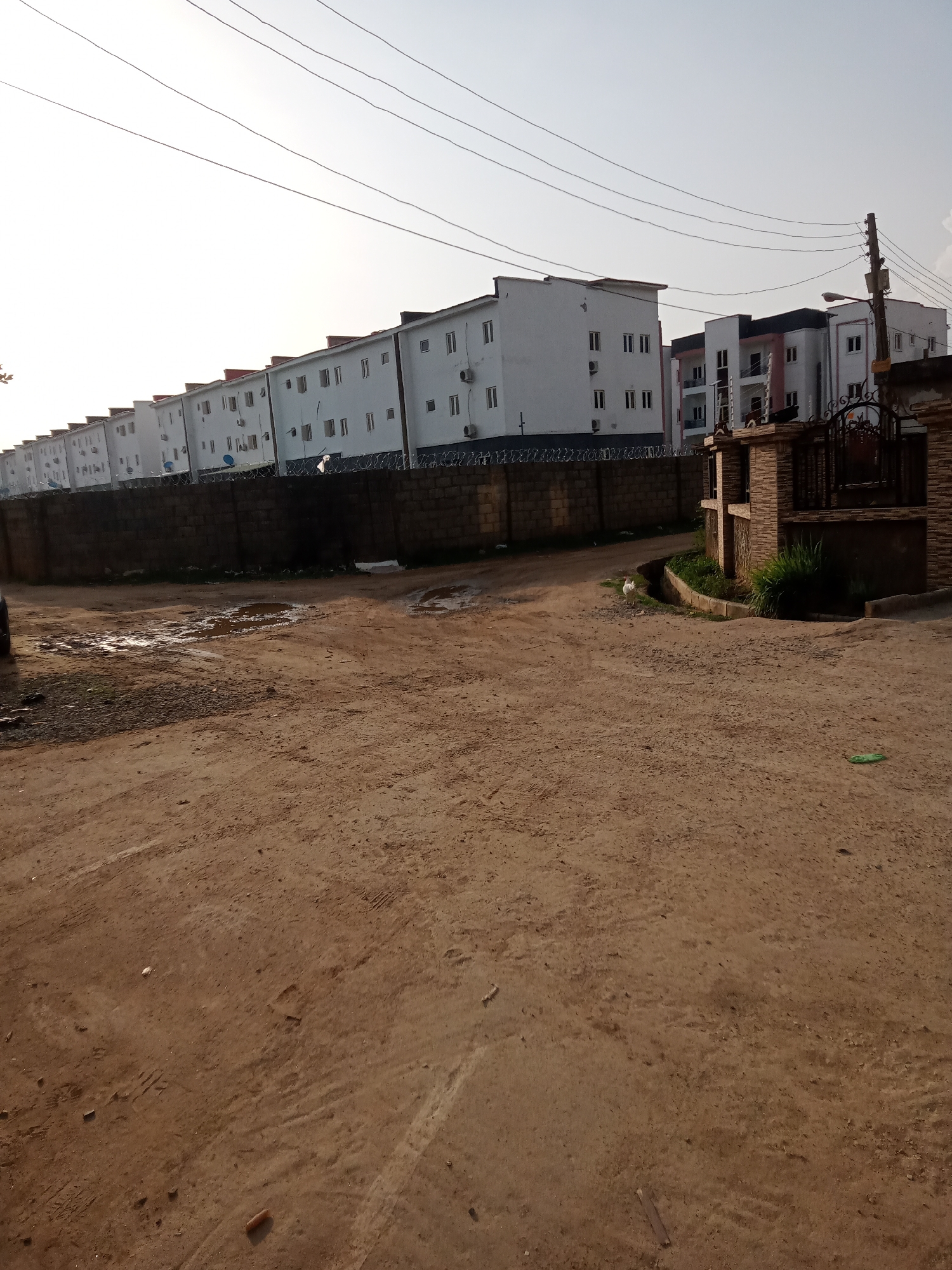 The scene depicts a dirt road in Gwarinpa, Nigeria, flanked by a long row of multi-story, white apartment buildings enclosed by a dark-colored brick wall topped with barbed wire. A drainage ditch runs along the side of the road, where a single white chicken stands near a gate. The road surface is uneven, with visible tire tracks, footprints, and small puddles. Overhead, electrical wires span across the sky. A utility pole is visible near the gate. The lighting suggests early morning or late afternoon.
