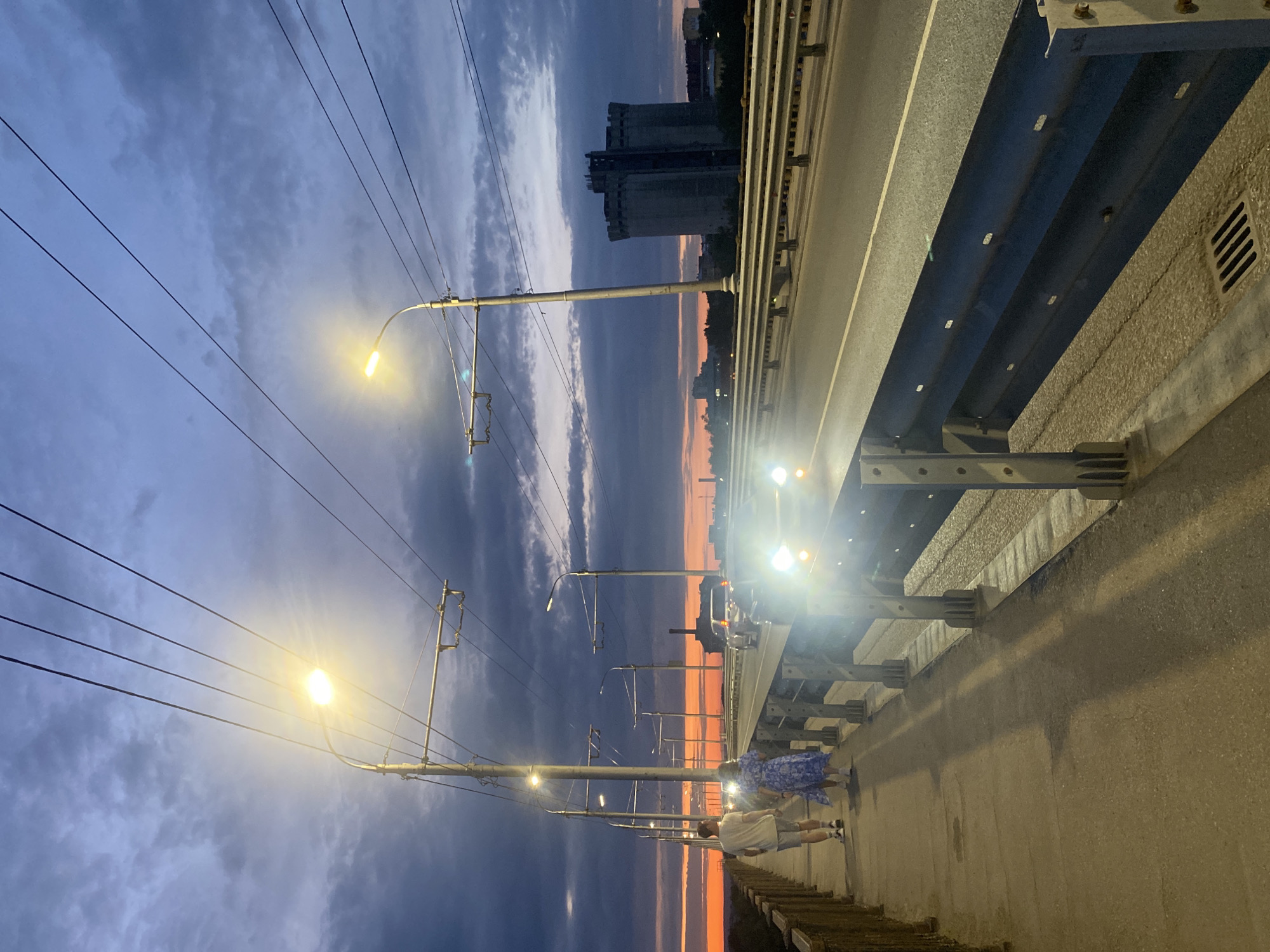 On a bridge in Novokuybyshevsk, Russia, during twilight, two individuals walk along a pedestrian path next to a road. The sky displays a gradient of purple and orange hues, with scattered clouds. A vehicle's headlights illuminate the road, indicating it is traveling towards the viewer. Numerous streetlights and overhead power lines stretch across the scene, with a tall, multi-story building visible in the distance. The pedestrian path is bordered by a metal railing.