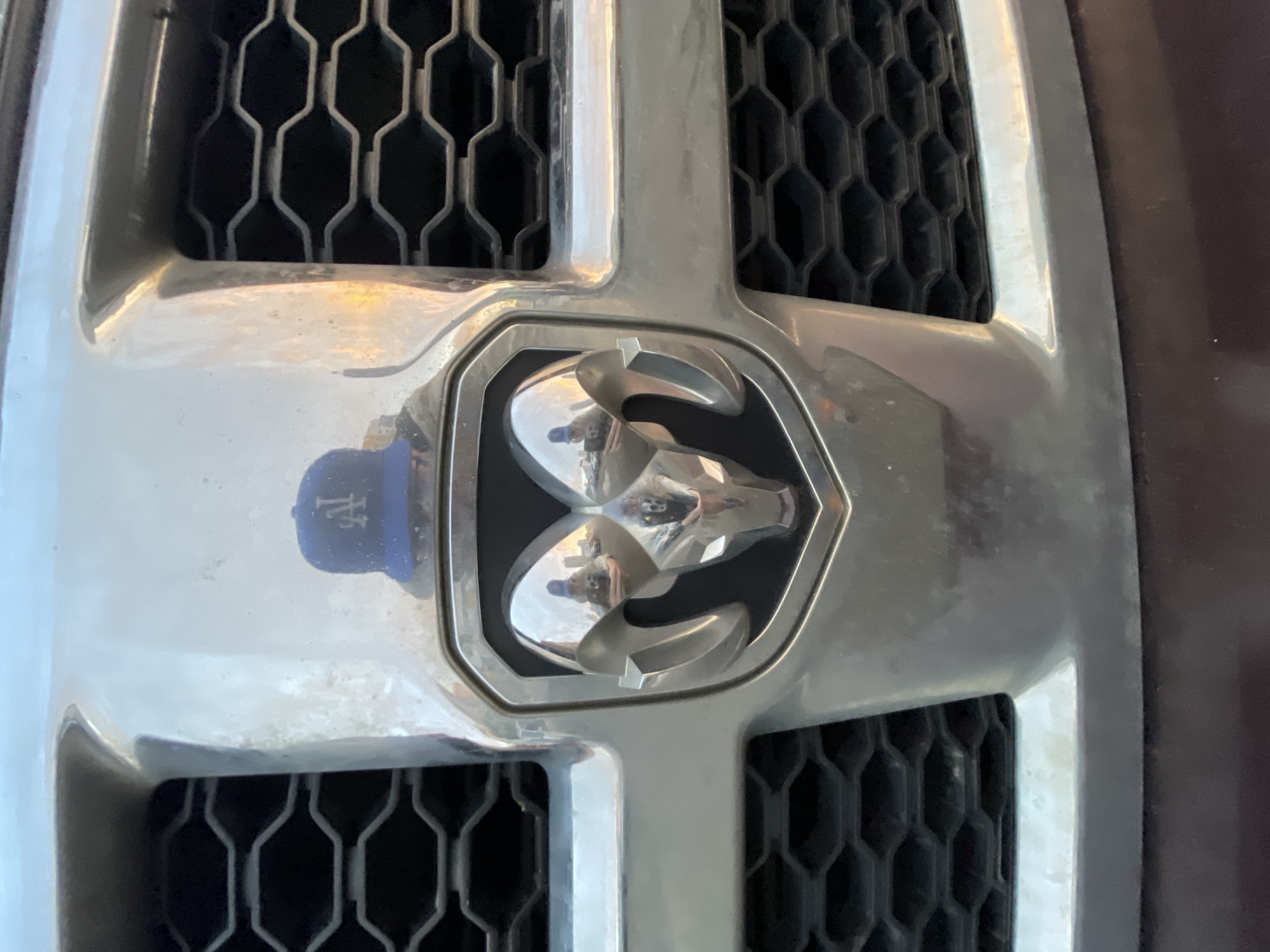 The media file is a close-up, slightly angled shot of the front grille of a vehicle. The central focus is the chrome emblem of a ram's head, which is set within a hexagonal frame. The emblem is highly reflective, showing distorted reflections of the surrounding environment. To the left of the ram's head emblem, a blue baseball cap with a white logo is partially visible, also reflected in the chrome. The grille itself features a honeycomb pattern. The overall tone is a bright, possibly daytime shot, given the lighting and reflections. There is no visible text on the vehicle's emblem or grille. The scene appears to be an outdoor setting, potentially a parking lot or driveway, though no specific location cues are evident beyond the vehicle's front end.
