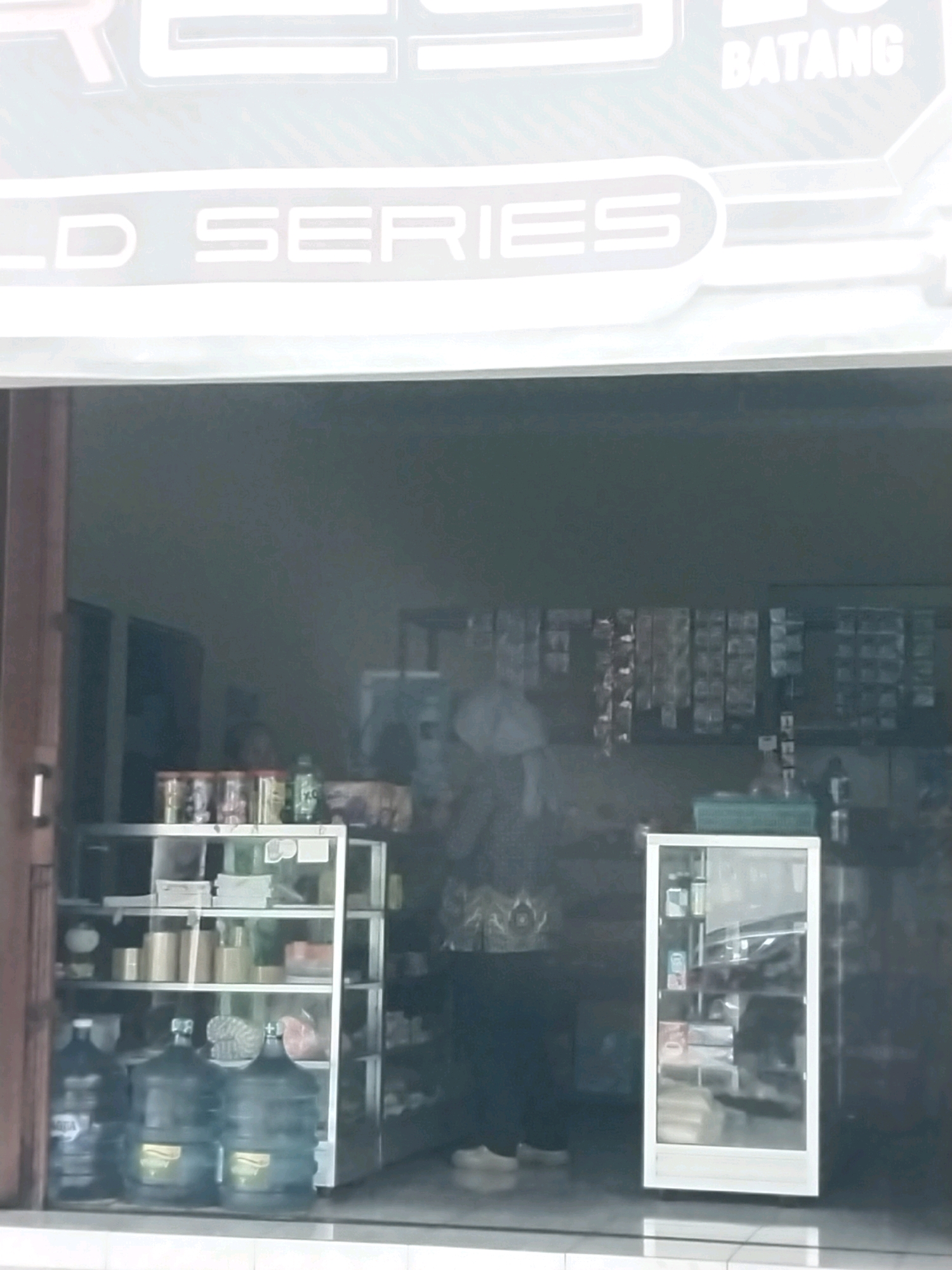 The image shows the exterior of a small shop with a glass storefront in Purwomartani, Indonesia. The shop appears to be a convenience store or a similar retail establishment.
Visible inside the shop are shelves stocked with various items, including what appear to be bottled drinks, packaged goods, and possibly snacks. There are several large water jugs on display near the entrance. A glass display cabinet is also present, showcasing more items.
The exterior of the shop features signage. One sign displays white text on a dark background, reading "LD SERIES". Another sign, partially visible at the top right, has the word "BATANG" in white text. The entrance to the shop has a sliding glass door. The floor in front of the shop is tiled.
The overall impression is of a typical, modest local shop in an urban or suburban Indonesian setting. The lighting suggests it is daytime, and the image quality indicates it might have been taken from a distance or with a low-resolution camera.