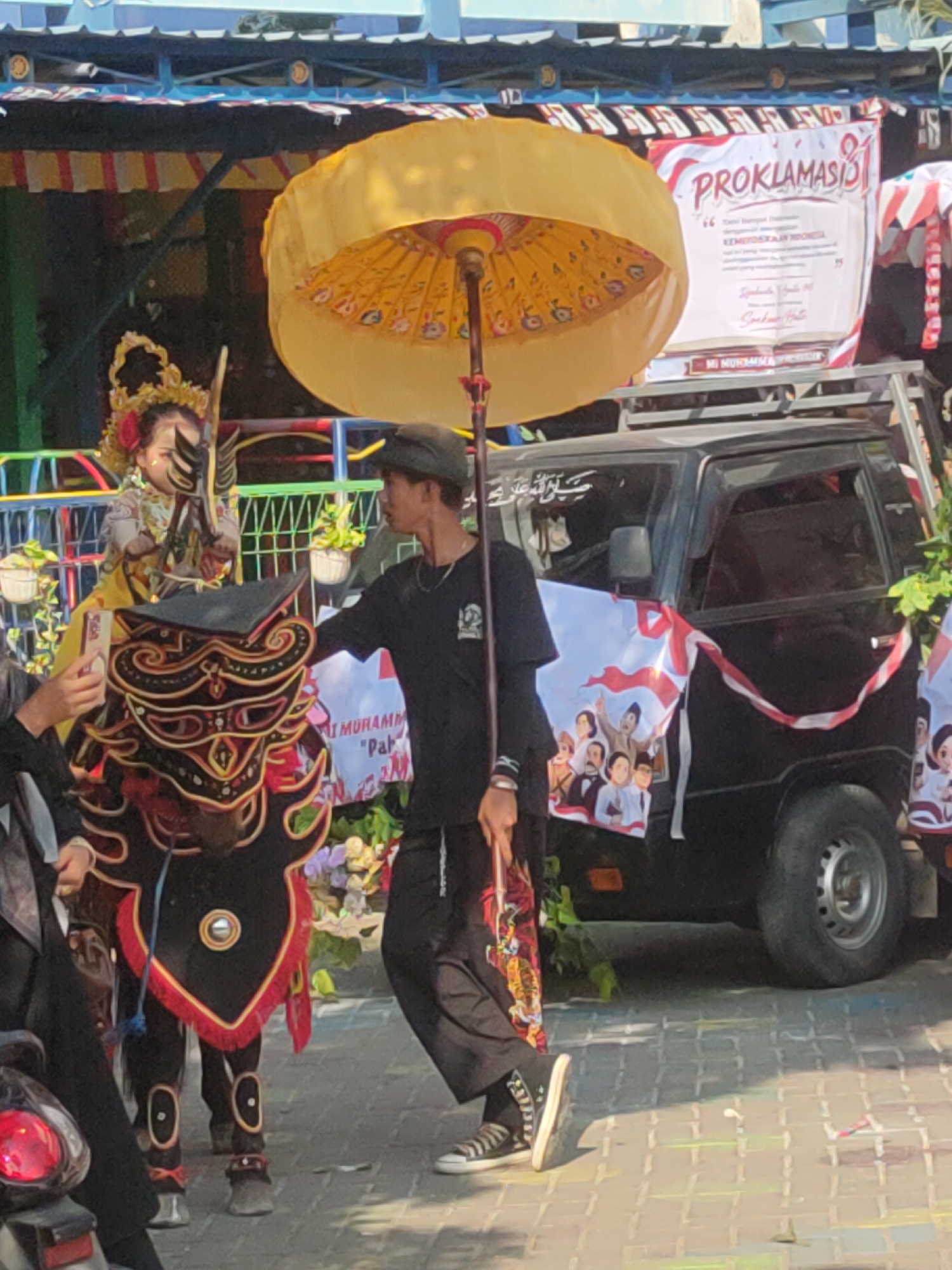 In the city of Payaman, Indonesia, a young person in traditional attire, possibly a dancer or performer, stands near a black van adorned with decorative banners. The van has a banner with the text "PROKLAMASI" in red and white lettering. Another banner on the side of the van features a group of people. A man in a black t-shirt and cap holds a large yellow umbrella over the performer, who is seated and wearing a headdress. The performer also has a decorative mask or costume piece in front of them, featuring intricate black, red, and gold patterns. The scene appears to be an outdoor event or parade, with a paved ground and a structure with a blue roof in the background. The overall atmosphere suggests a cultural celebration or festival.