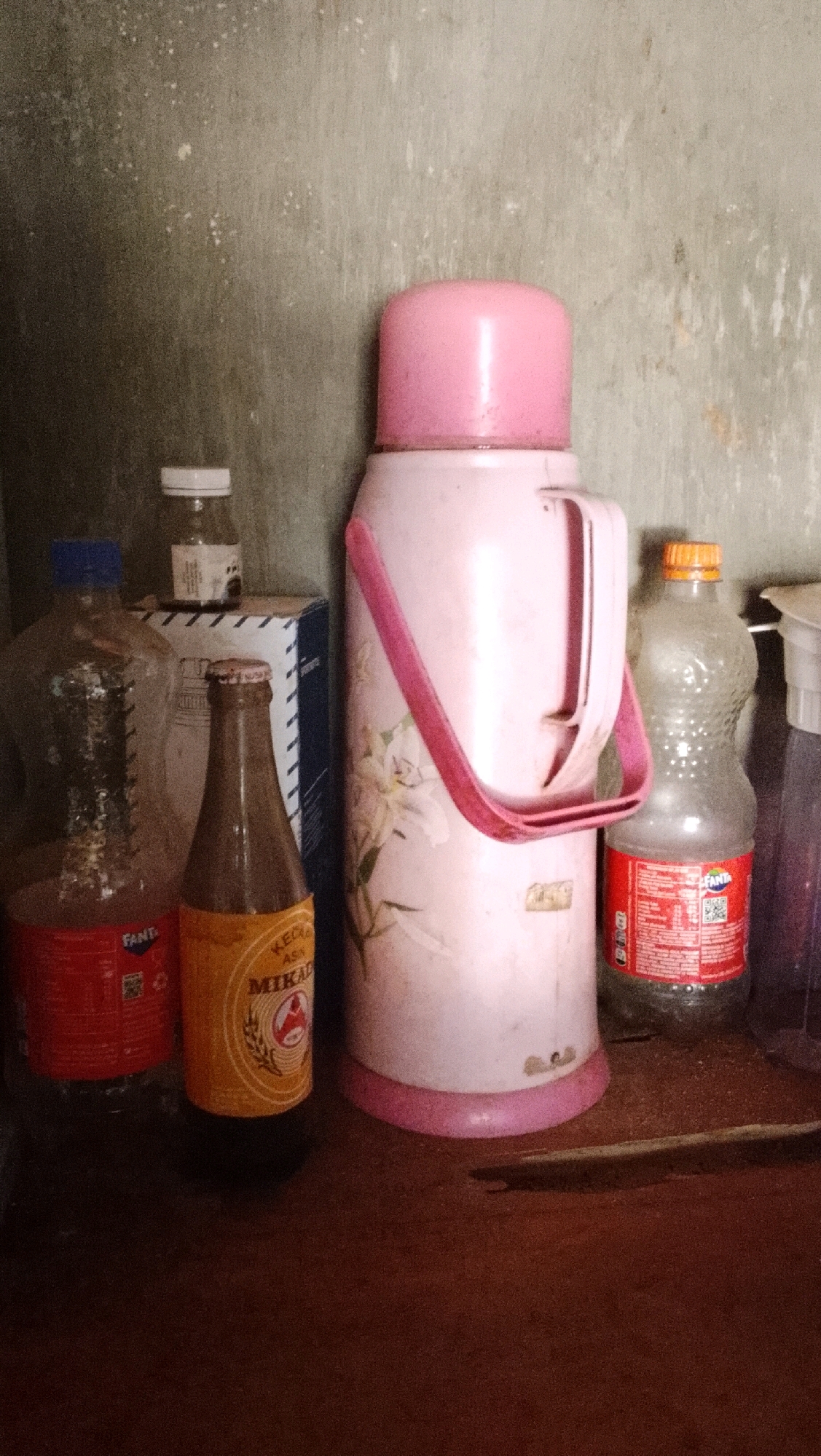 In Bojong Barat, Indonesia, a collection of household items is arranged on a wooden surface against a textured, grey wall. The scene features a large, pale pink thermos with a pink strap, adorned with a floral design. To its left, a clear plastic bottle with a red label, appearing to be a Fanta bottle, stands beside a smaller brown glass bottle with a yellow label that reads "MIKAD". Behind these bottles, a blue and white box is partially visible. To the right of the thermos, another clear plastic bottle with an orange cap and a red label, also resembling a Fanta bottle, is present. A small white bottle with a white cap sits behind the box, and a partially obscured purple container is on the far right. The lighting suggests an indoor environment, possibly a kitchen or storage area.