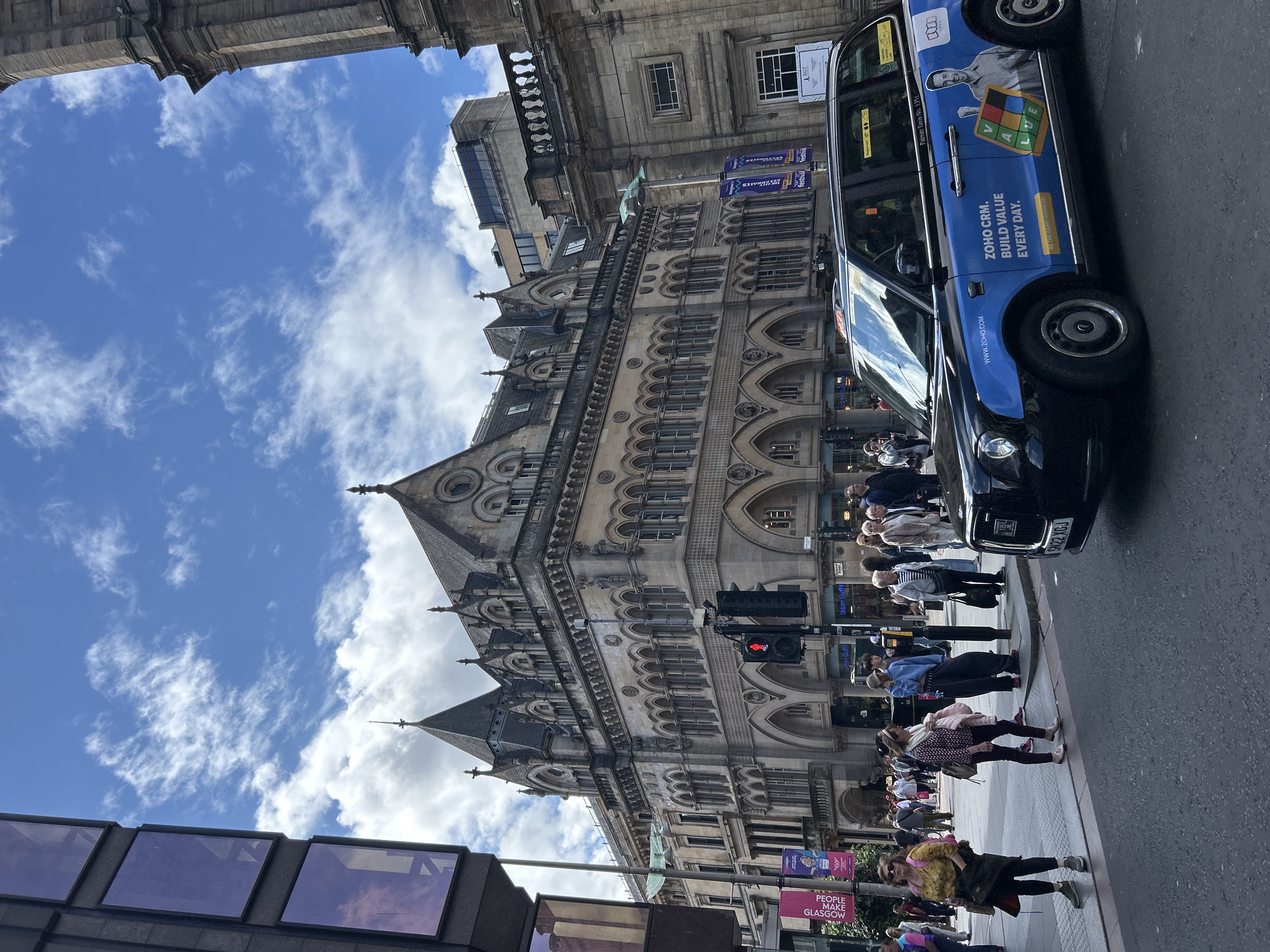An eye-level shot of a busy street scene in Glasgow, United Kingdom, on a partly cloudy day. A black taxi, adorned with a bright blue advertisement for "ZOHO CRM," is parked in the foreground, its passenger side facing the viewer. The advertisement features text like "BUILD VALUE EVERY DAY" and an image of a smiling man holding a Scrabble-like tile arrangement spelling "VALUE." The taxi has a license plate that reads "SN22 XGJ."
Behind the taxi, a diverse group of pedestrians are waiting at a street corner. A woman in a yellow top and sunglasses stands prominently on the left, holding a dark bag. Next to her, a woman in a patterned top and dark pants carries a pink jacket over her arm. Other individuals are visible in the background, some standing on the pavement, others crossing the street.
The urban environment is dominated by impressive architecture. A grand, ornate stone building with gothic and Victorian elements stands tall, featuring numerous arched windows and intricate detailing. Adjacent to it is another stone building, simpler in design. A tall traffic light with a red signal is visible. Banners with the text "PEOPLE MAKE GLASGOW" and "festival" hang from lampposts, indicating a possible event or cultural celebration. The sky above is a vibrant blue, streaked with white, wispy clouds.