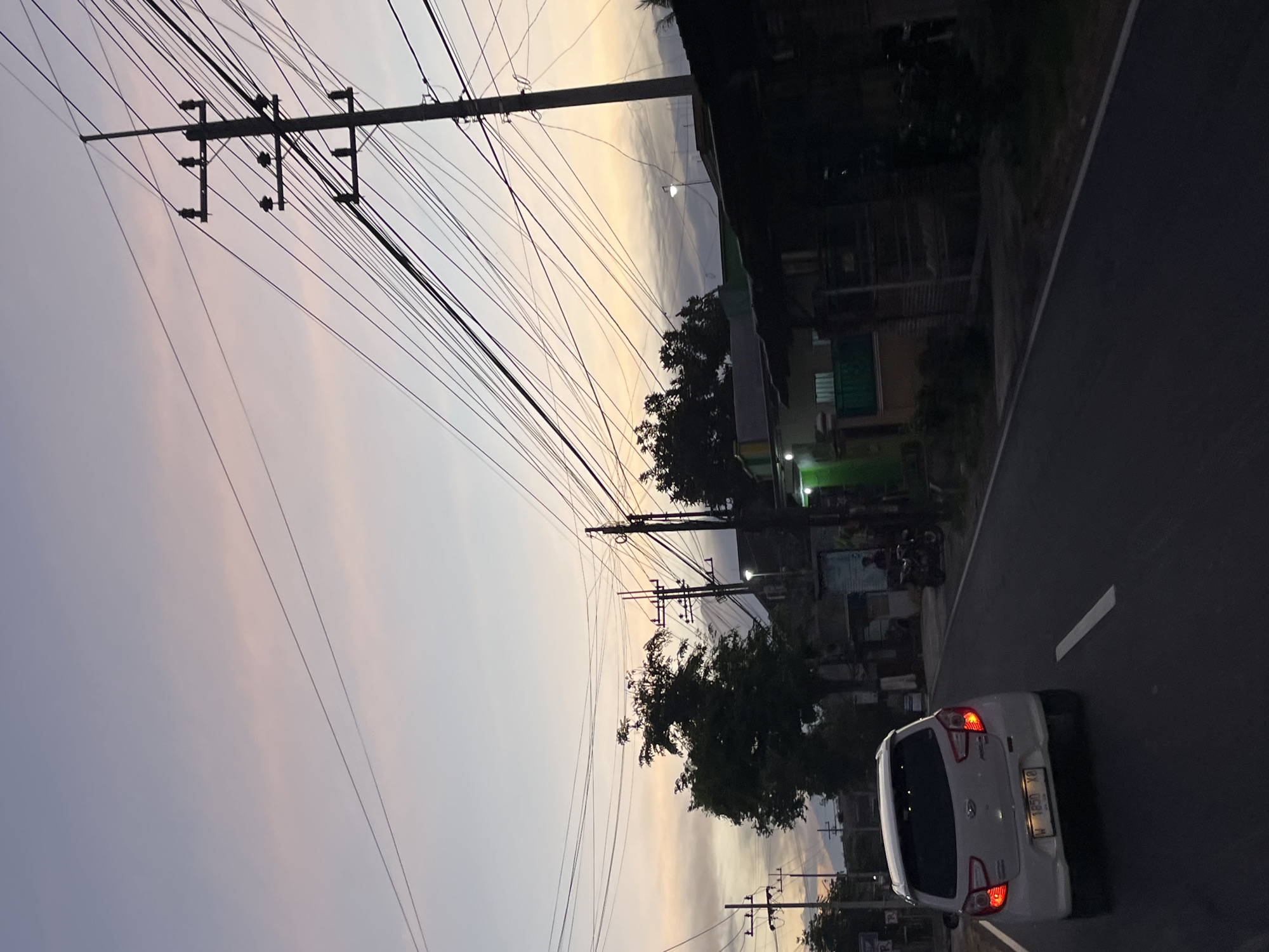 This media file captures a street scene at dusk in Purworejo, Indonesia. The image is oriented vertically, with the sky occupying the left portion and the road and buildings on the right. A white car with its taillights illuminated is driving away from the viewer on the right side of the road. The car's license plate is visible and reads "W 1850 YP". Overhead, a dense network of power lines crisscrosses the sky, supported by utility poles. The sky itself displays a gradient of colors, transitioning from a soft purple and pink near the horizon to a pale yellow and gray higher up, indicating the twilight hour.
On the right side of the road, buildings and trees are visible, partially obscured by the dim light. Some of the buildings appear to be residences or small shops, with some lights on inside. A motorcycle is parked in front of one of the buildings. The overall atmosphere is calm and quiet, typical of the early evening in a residential or semi-urban area. The presence of the power lines and the car suggests a functional, inhabited location. The weather appears to be clear, allowing for the visibility of the sunset colors.