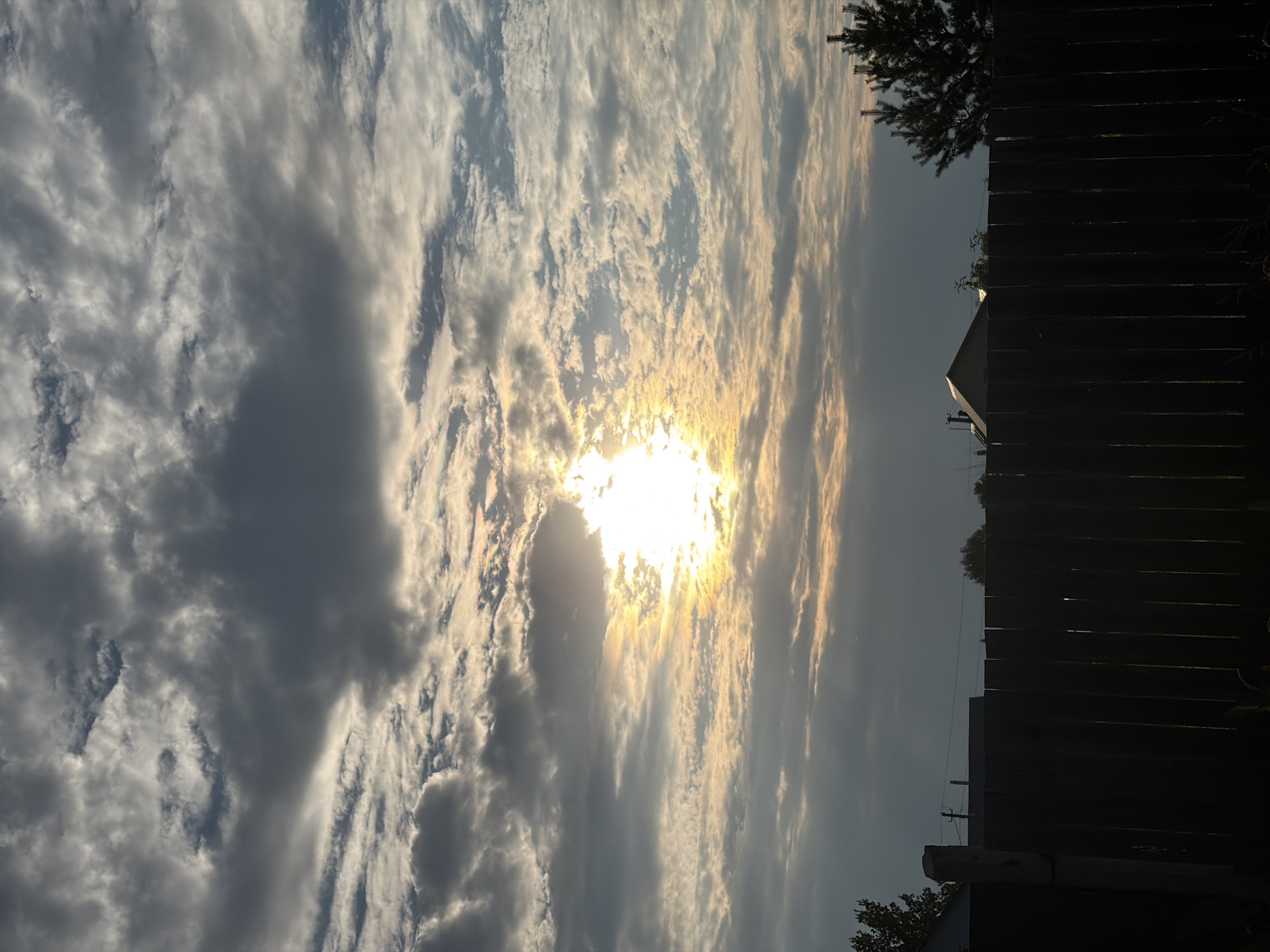 The image shows a view of the sky over a residential area, likely in the village of Olkhovka, Russia, during sunset. The sky is filled with a mix of dark grey and white clouds, with the sun visible behind the clouds, casting a bright, warm glow. The lower portion of the image is dominated by a dark, wooden fence that runs horizontally across the frame. Behind the fence, the silhouettes of several houses and a pine tree are visible against the darkening sky. The scene suggests a rural or suburban setting.