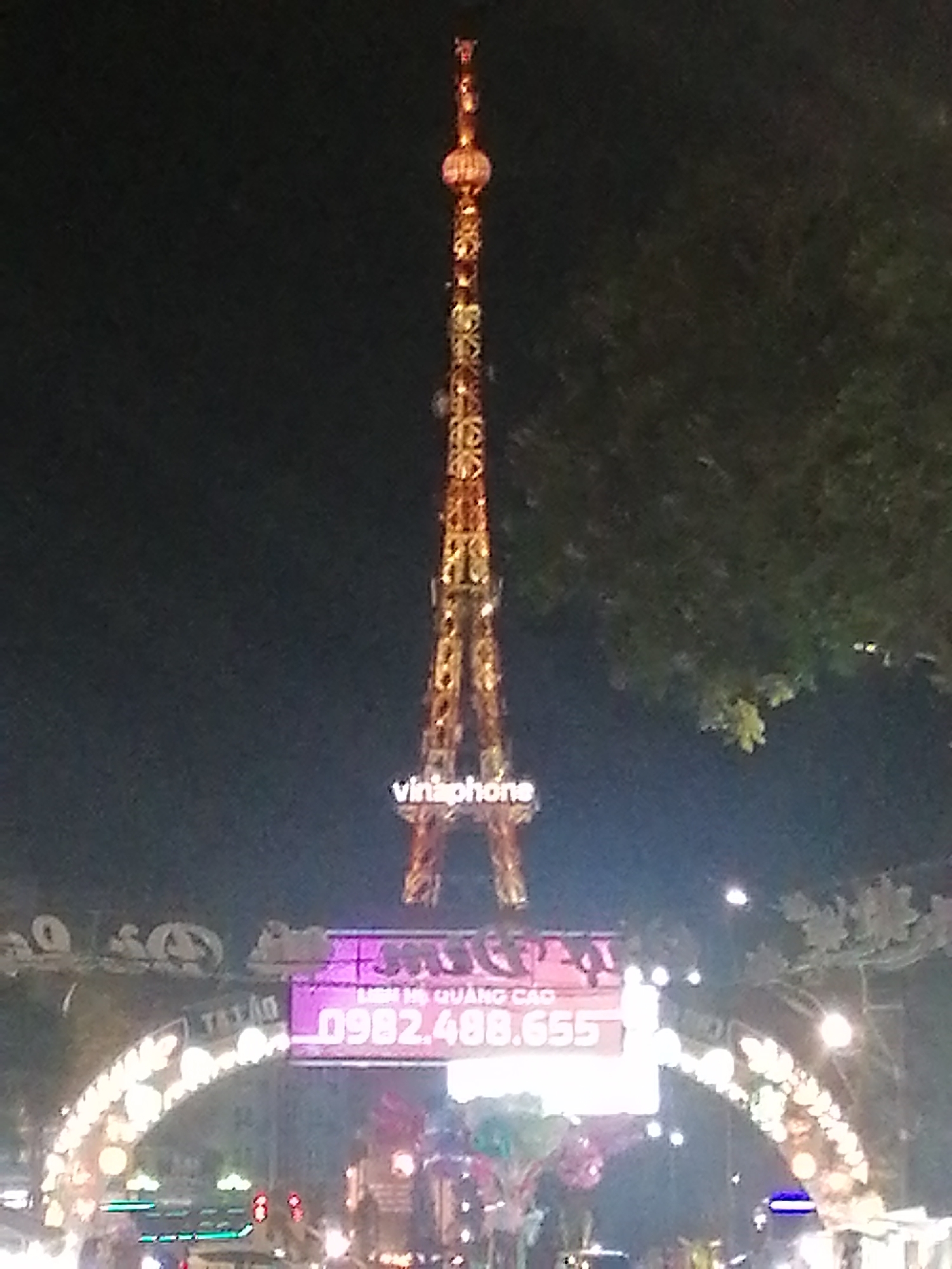 At night in Da Lat, Vietnam, a tall communications tower stands above a decorated city gate. The tower is lit with orange lights and displays the word "vinaphone" in illuminated letters near its base. Below the tower, the city gate is adorned with strings of lights and a digital display showing a phone number. Vehicles and pedestrians can be seen in the blurred foreground, suggesting a bustling urban environment. The sky is dark, and trees are visible on the right side of the frame, indicating the presence of greenery within the city.