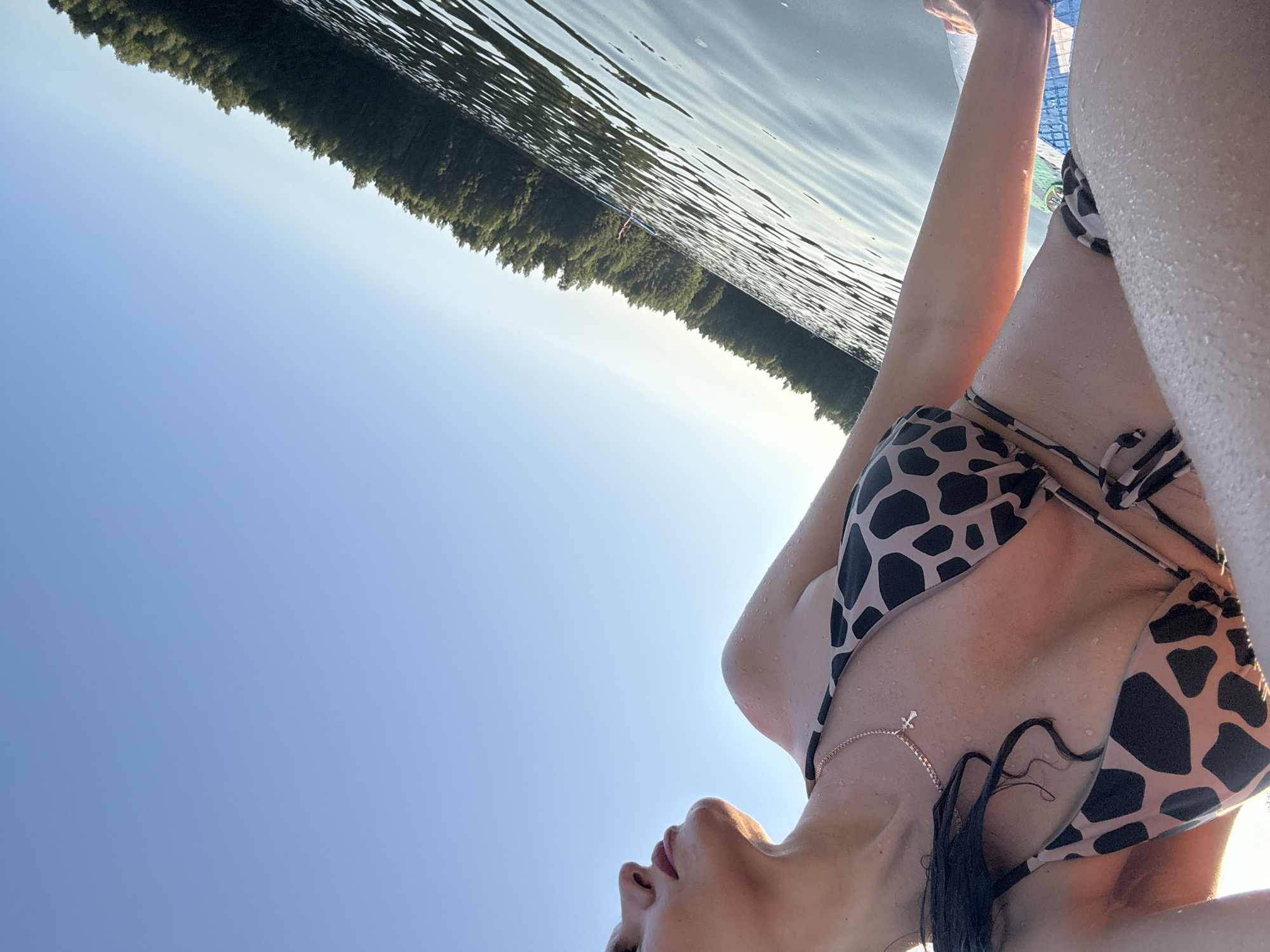 A low-angle, full-frame, eye-level shot captures a woman in a bikini on a body of water, likely in Chernushka, Russia. The woman's upper torso and right arm are visible, with her head turned away from the camera and a profile of her face showing. She is wearing a tan bikini with black spots and a delicate silver necklace with a cross pendant. Her skin is wet, with water droplets visible on her arm and chest. The background consists of a clear blue sky and a distant treeline bordering the water. The water is calm with gentle ripples. A person on a paddleboard can be seen in the distance. The lighting suggests it is late afternoon or early evening, with the sun casting a warm glow on the woman's skin and the water.
