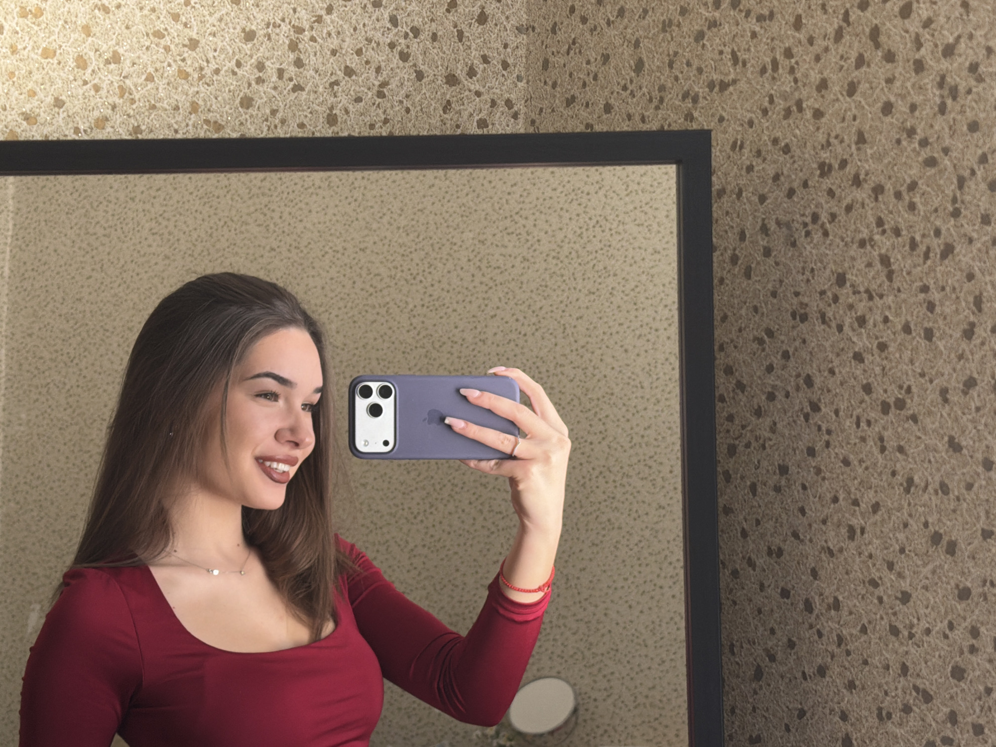 A young adult female is depicted taking a mirror selfie in an indoor setting, identified as a location within Sofia, Bulgaria. The subject, with long brown hair, is wearing a dark red, long-sleeved top with a scoop neckline and a delicate silver-toned necklace. She is smiling, looking towards her left, and holds a purple-cased smartphone horizontally with her right hand. Her right wrist displays a thin red beaded bracelet, and her fingernails are painted light pink. A ring is visible on her right ring finger. The smartphone, identifiable as an iPhone with a triple-lens camera array, is pointed towards her reflection in a dark-framed rectangular mirror. The wall behind the mirror exhibits a textured, mottled pattern in shades of beige and brown with darker irregular spots. A smaller, circular, stand-mounted mirror is partially visible within the main mirror's reflection in the lower right.