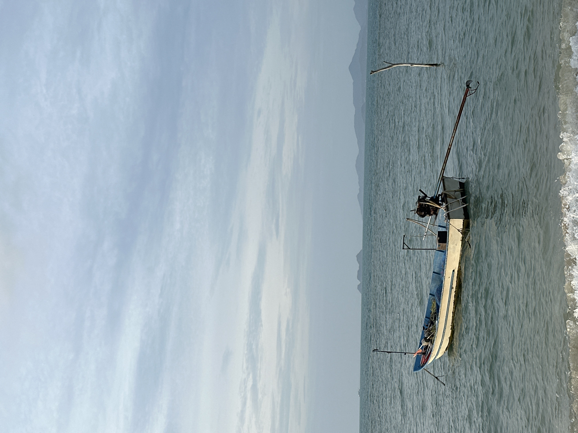 A longtail boat is anchored in calm, blue ocean water, near the shore of Ko Samui District, Thailand. The boat is white with a blue stripe and has fishing equipment and a motor attached. In the distance, several islands are visible on the horizon under a cloudy sky. A dead tree stands in the water to the right of the boat, and small waves crash on the sandy shore in the foreground.
