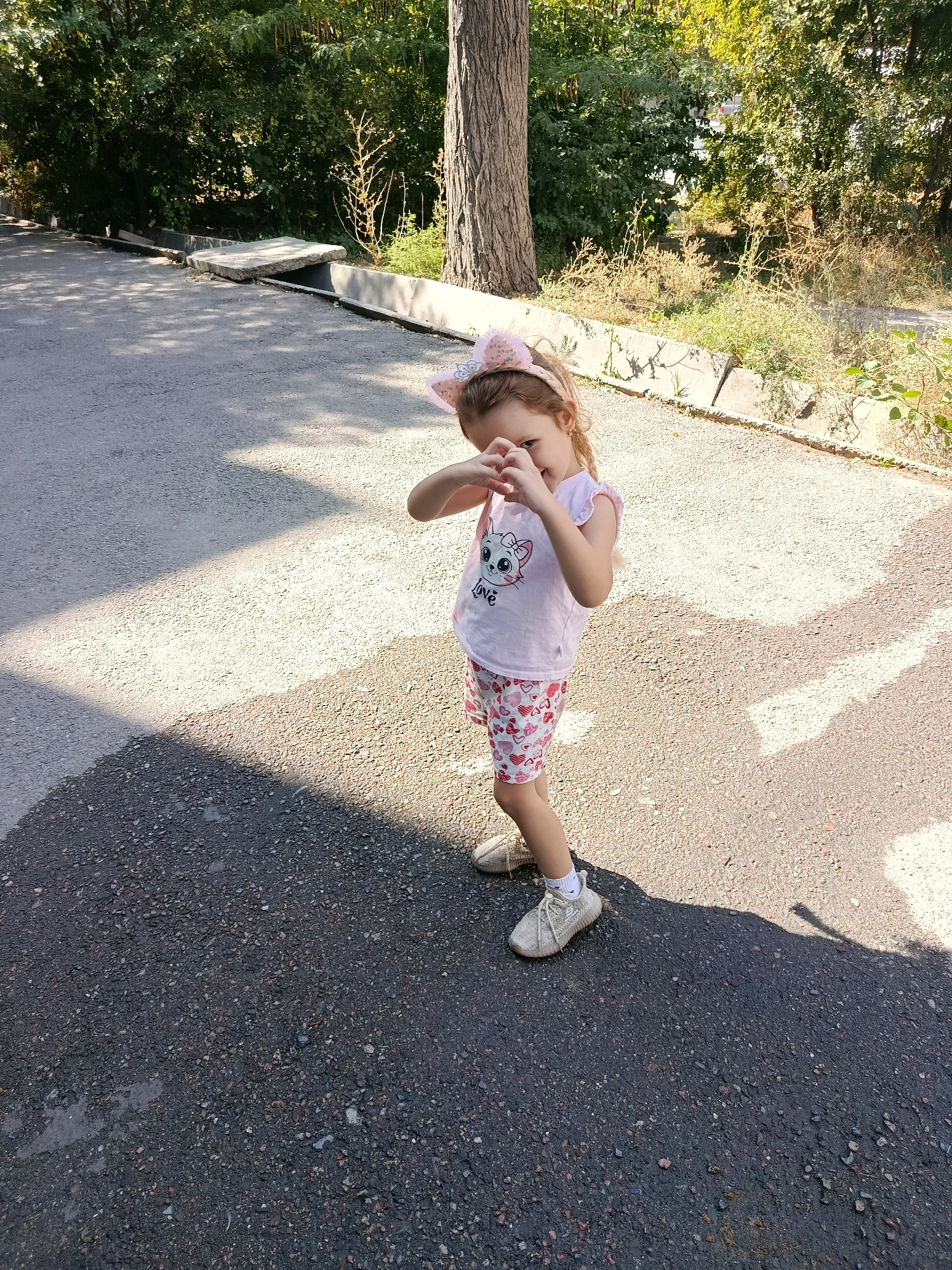 A young girl with blonde hair and a pink headband with cat ears stands on an asphalt pathway. She is wearing a pink t-shirt with a cat graphic and white text, and pink shorts with a heart pattern. Her hands are held up in front of her face, forming a heart shape with her fingers. She is wearing light-colored sneakers. The pathway is partially shaded, with a large shadow cast from the left side of the frame. In the background, there are trees, bushes, and a concrete structure that appears to be a curb or low wall. The scene appears to be outdoors, possibly in a park or on a sidewalk in Bishkek City, Kyrgyzstan.