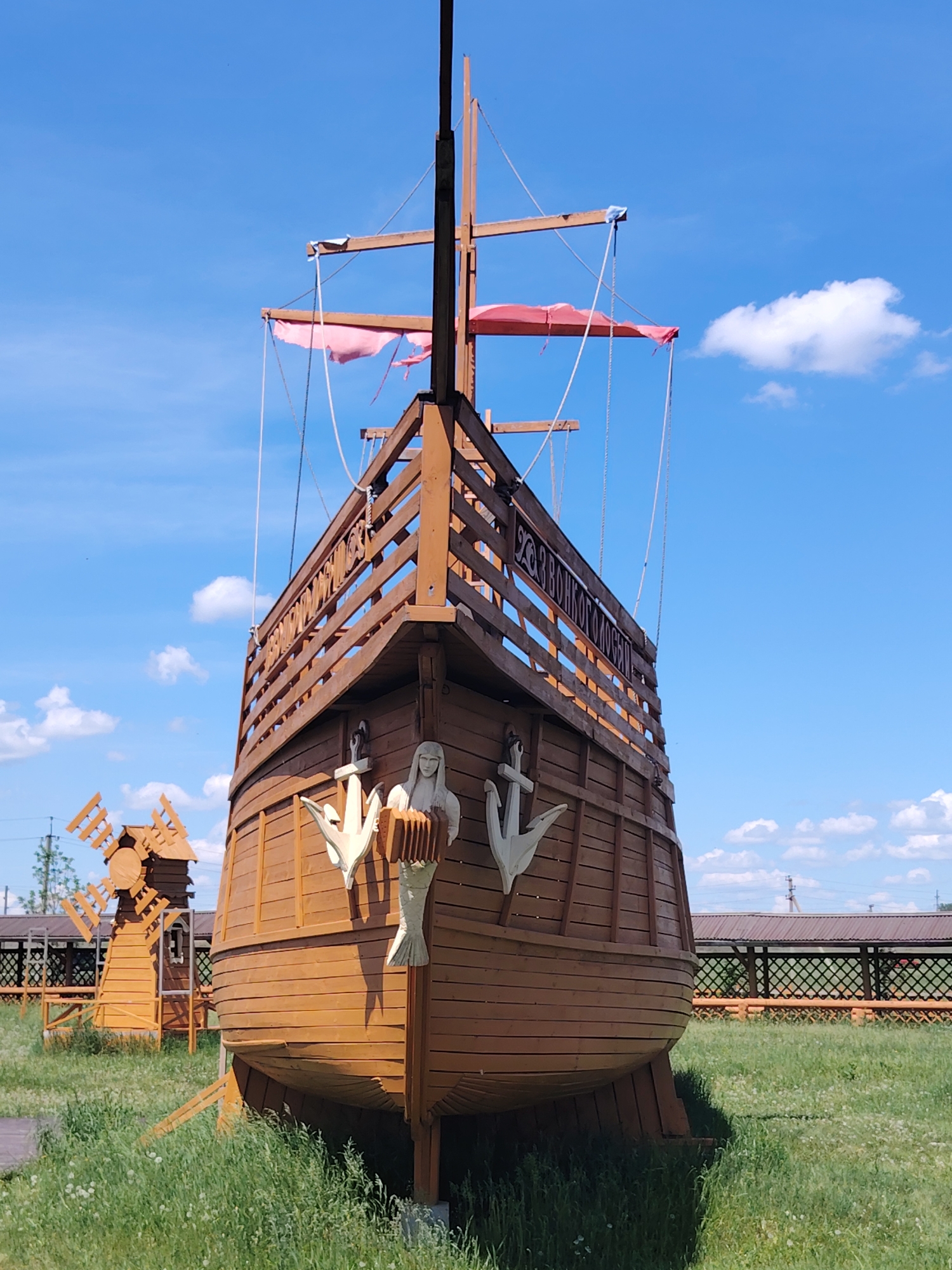 An outdoor, eye-level, full shot captures a wooden replica of a ship with red tattered sails against a bright blue sky with scattered white clouds. The ship's bow points directly towards the viewer, revealing detailed wooden planking and decorative elements. To the left of the ship, a small, wooden windmill is visible. The scene is set in a grassy field with a wooden fence and buildings in the background. The ship's hull is adorned with two white anchors and two mermaid-like figures holding musical instruments. Cyrillic text is visible on the ship's railing.