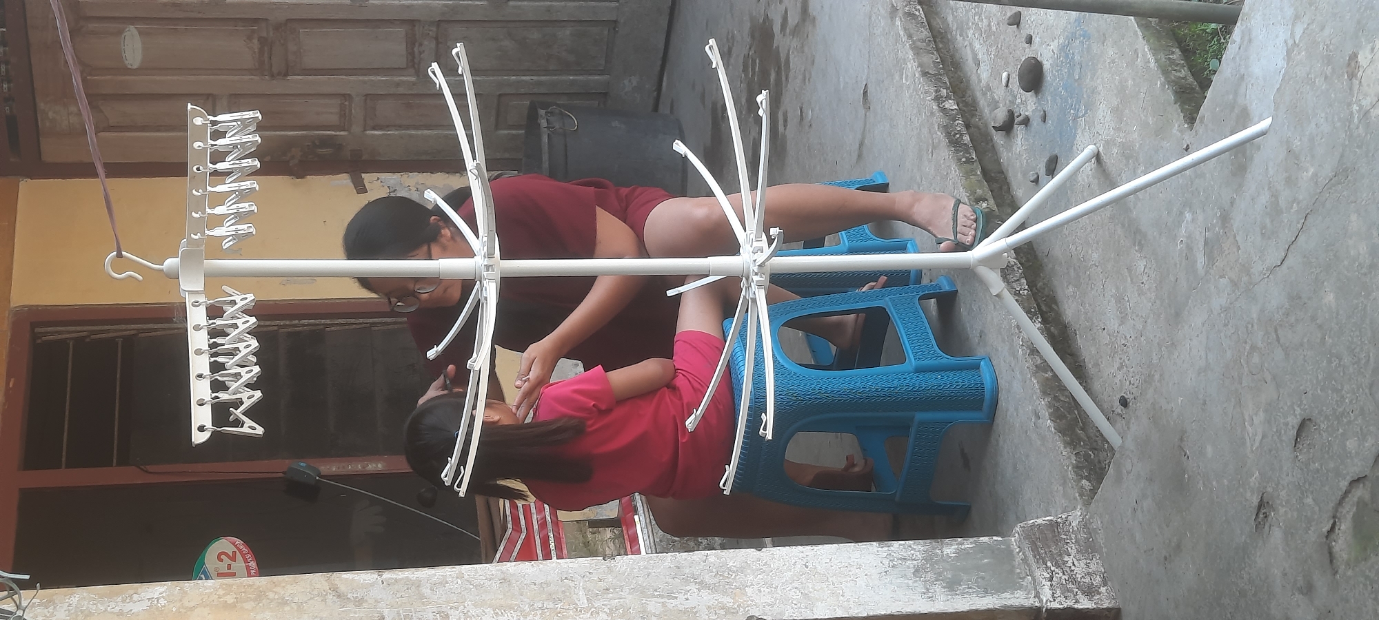 In Ploso, Indonesia, a person with glasses, wearing a maroon shirt, is cutting the hair of a young child. The child, wearing a pink t-shirt and shorts, is seated on a blue plastic stool. They are positioned in front of a white, multi-tiered drying rack. The scene takes place outdoors on a concrete surface, with a weathered building and a wooden door visible in the background.