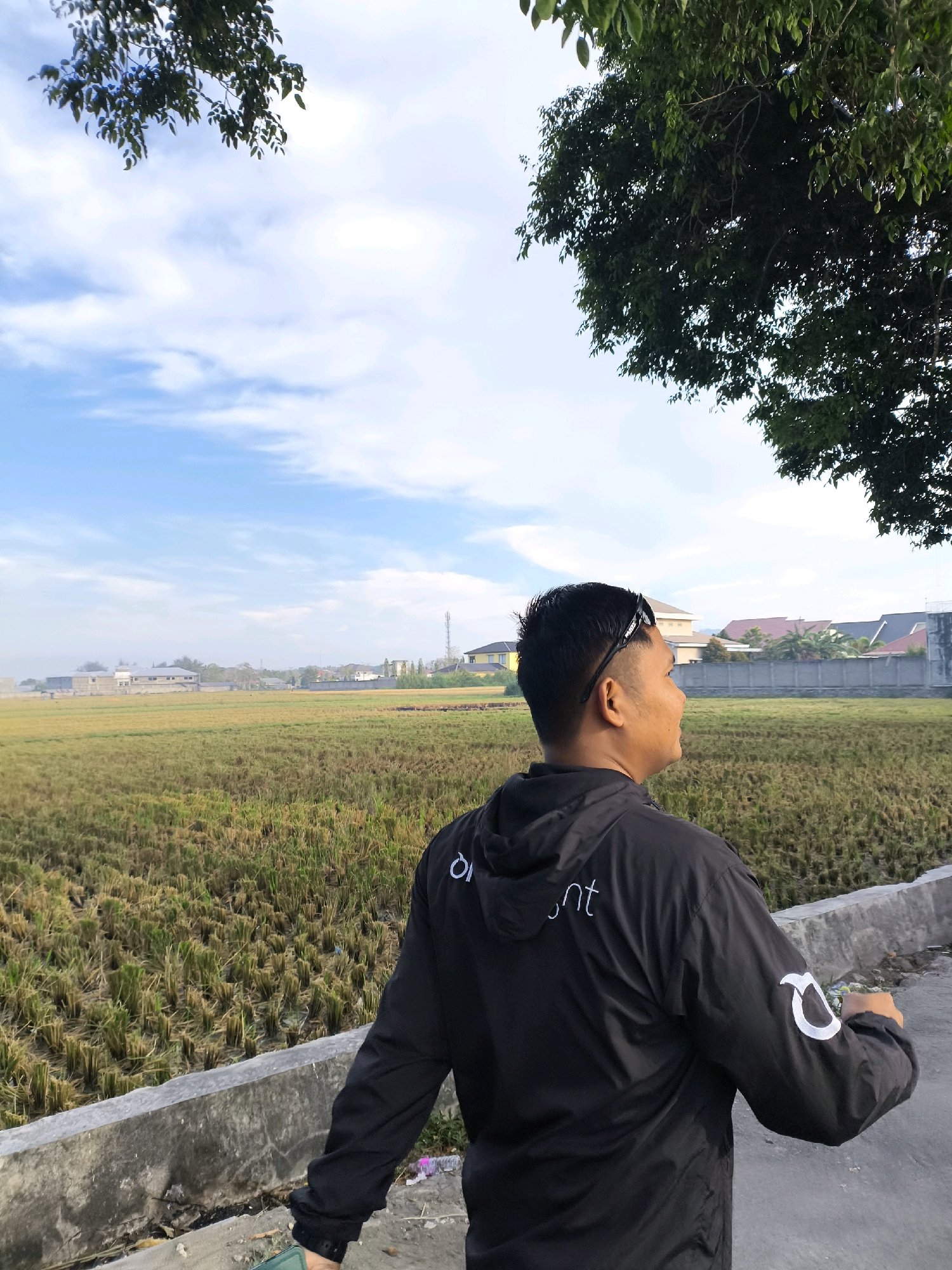 A man wearing a black jacket with white lettering is standing next to a rice field under a partly cloudy blue sky. He is looking to the right, away from the camera. The rice field is dry and appears to have been harvested. In the background, there are buildings and a cell tower, suggesting an urban or suburban environment. The scene is set in the city of Gorontalo, Indonesia.