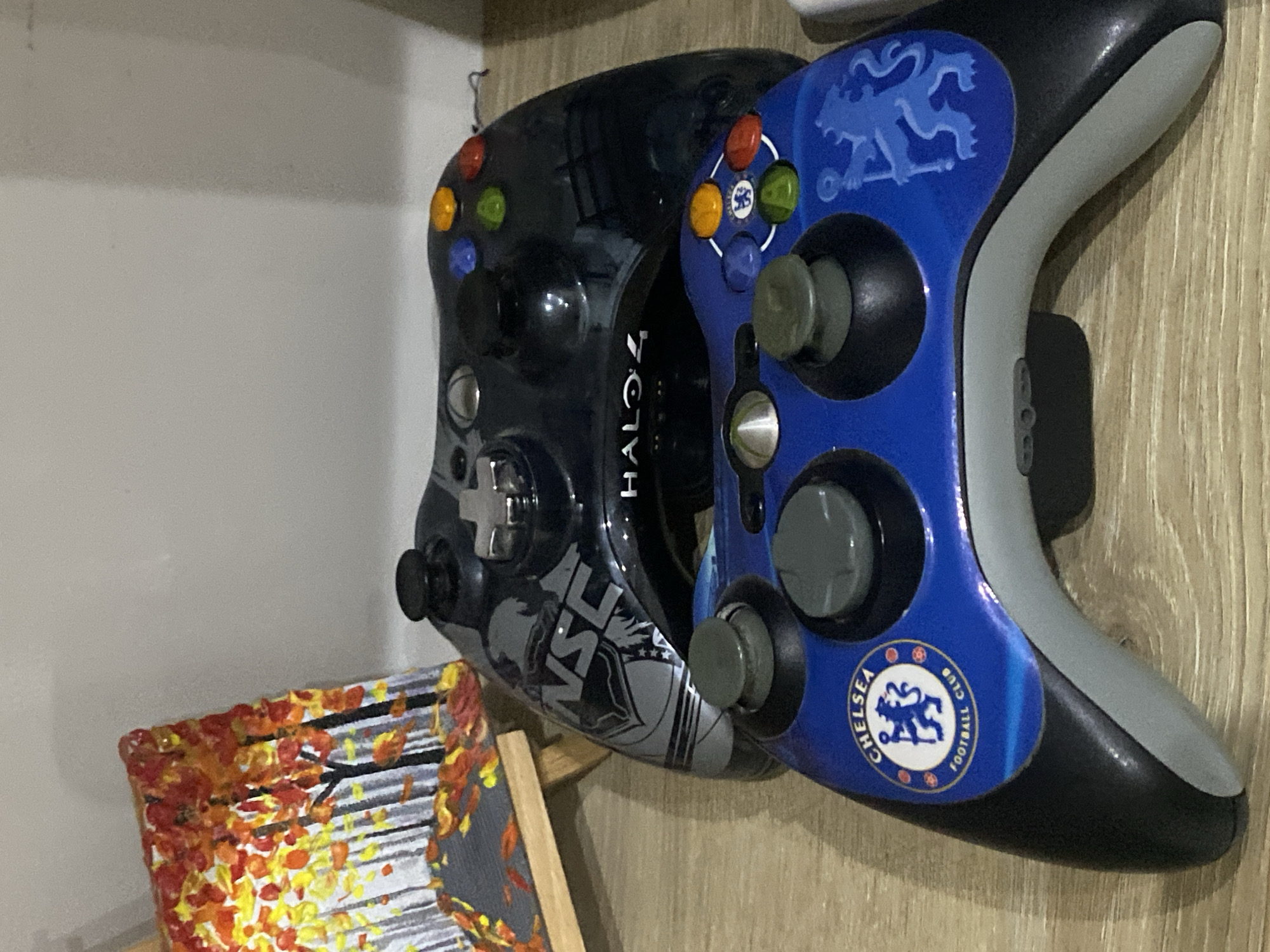 Two video game controllers are stacked on a wooden surface. The top controller is black with "Halo 4" branding and a gray design featuring a knight's helmet. The bottom controller is blue with the Chelsea Football Club logo and a stylized blue lion. In the background, a small painting of autumn trees on an easel is visible. The scene appears to be indoors, likely in a room. There is no indication of the specific location within Chișinău, Moldova.