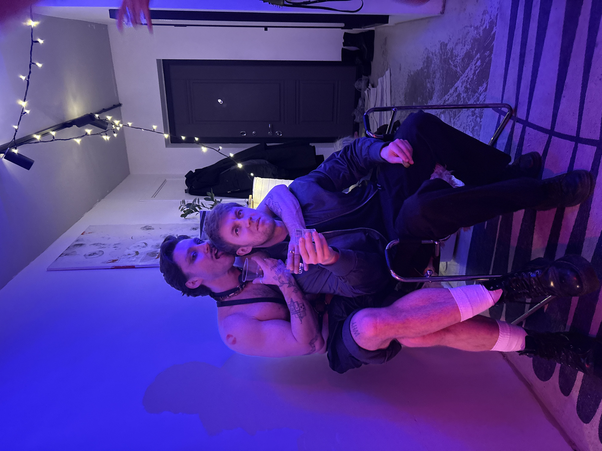 Two men are close together in an indoor setting, illuminated by colored lights. One man is shirtless with a dark collar, tattoos on his arm, and a red mark on his chest. The other man is wearing a jacket. Both are holding small glasses. They are seated in a metal chair. String lights are visible. Behind them is a dark door and what appears to be a piece of art. The location is likely not Moscow, Russia, as there is no evidence of this in the image.