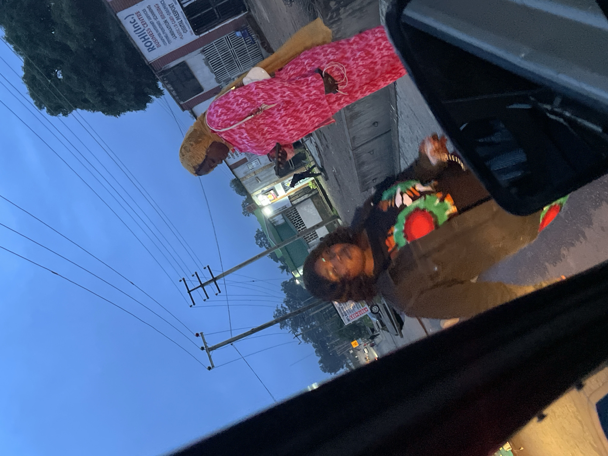 In Jos, Nigeria, two women stand on a street at dusk. The woman on the right wears a bright pink patterned dress and a yellow headscarf, holding a phone. To her left, a woman in a dark jacket with a colorful floral design on her shirt looks towards the camera. Behind them, a building displays a sign for "ROHI(Inc.) BUSINESS CENTRE" listing services like computer/internet services, typing, printing, scanning, lamination, building, plastic ID card graphics, and digital passport. Utility poles with power lines are visible in the background, and the sky is a deep blue. The foreground is occupied by the dark interior of a vehicle, possibly a car, with a side mirror reflecting a portion of the scene. The overall atmosphere suggests an urban setting during twilight hours.
