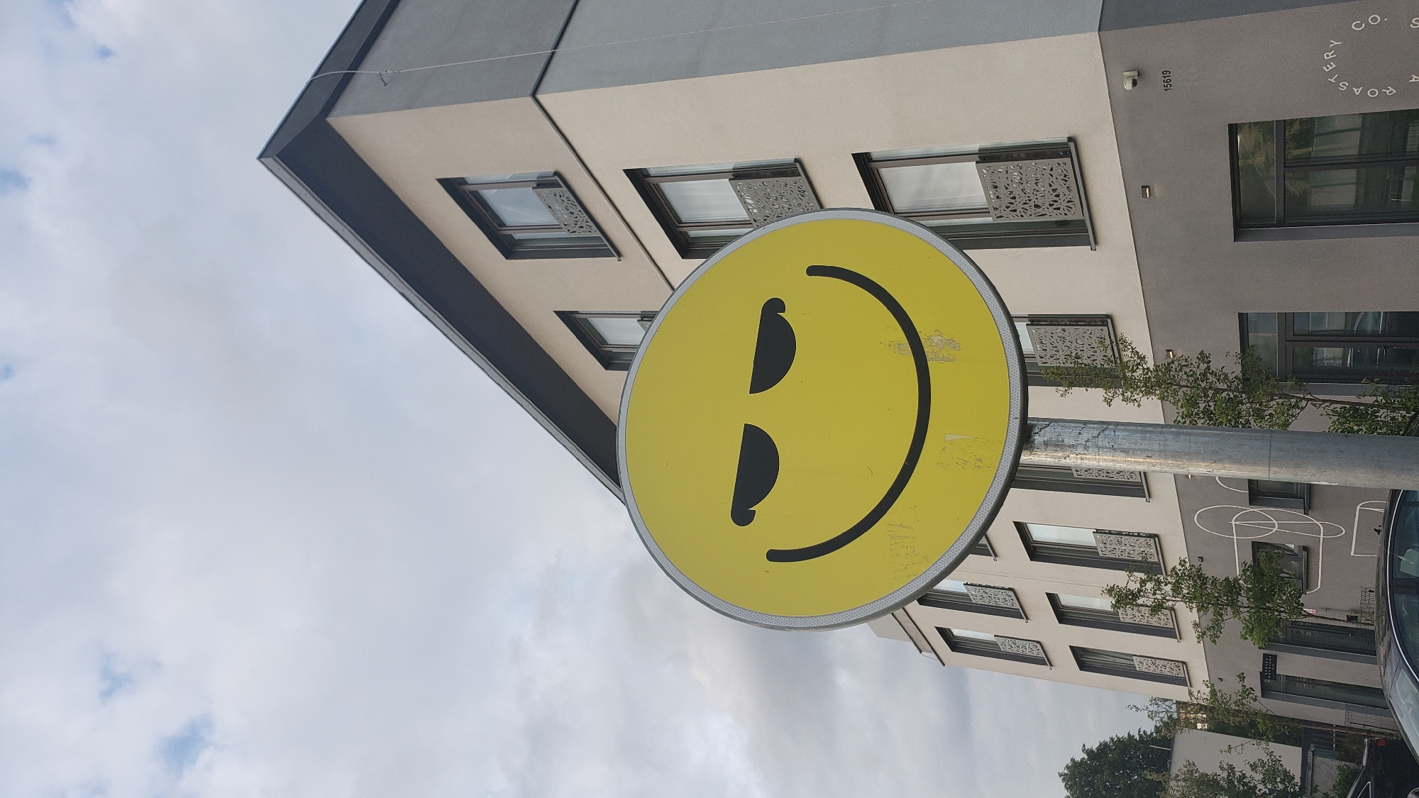 A slightly angled, medium shot shows a yellow smiley face sign on a silver pole. The sign is positioned in front of a multi-story building with multiple windows. Some of the windows feature decorative metal panels. A small tree and part of a vehicle are visible near the bottom right. The sky is cloudy. The sign is located in Prešov, Slovakia, specifically near a branch of the KIERI ROASTERY. The building behind the sign has the number 15619.