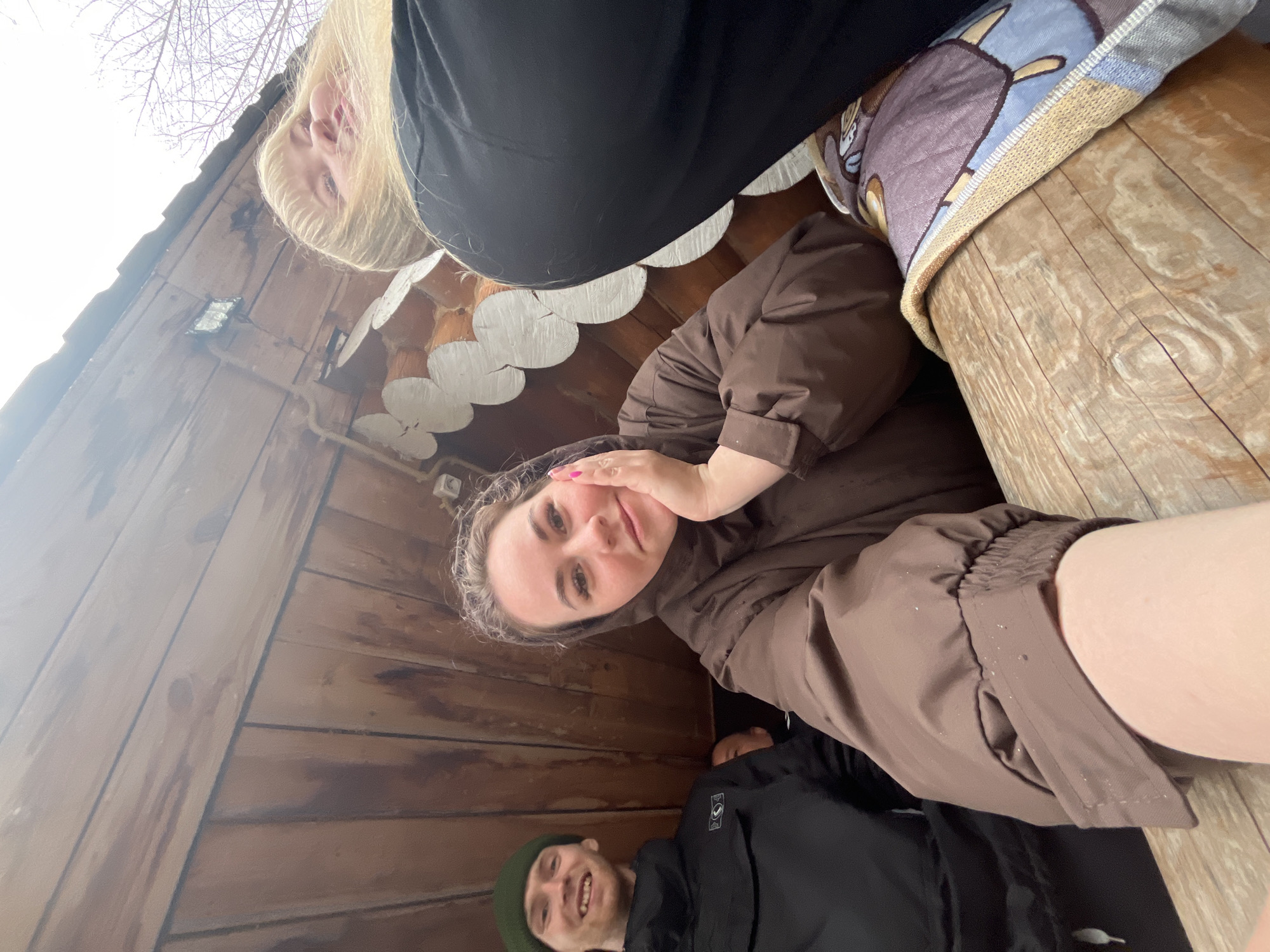 Three individuals are visible in the image, taken from a low-angle perspective, possibly within a wooden structure. A fair-skinned woman with blonde hair is positioned at the top of the frame. Below her is a fair-skinned woman with brown hair, wearing a brown jacket, and holding her hand near her face. At the bottom of the frame, a man with fair skin is smiling, wearing a green beanie and a black jacket. A log-like object is behind the brown-haired woman, and a pillow is resting on it. The interior walls of the structure are made of wood. The location is Chaikovsky, Russia.