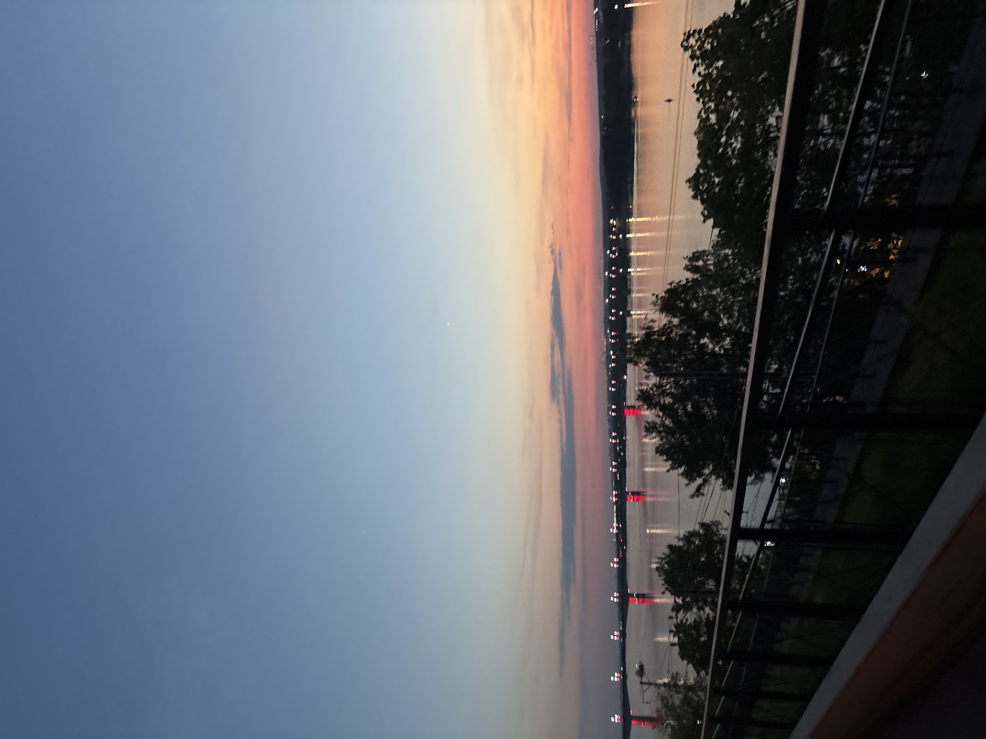 The image depicts a waterfront scene in Perm, Russia, at dusk. A body of water reflects the red and white lights along the shoreline, possibly from bridges or buildings. Trees and a modern railing are visible in the foreground, suggesting the view is from an elevated platform or balcony. The sky transitions from blue to pink and orange hues, indicating either sunrise or sunset. No people are visible in the frame.