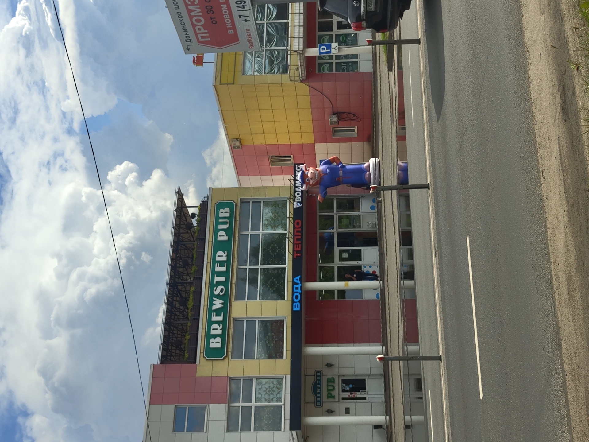 The image shows a street scene in Dergayevo, Russia, with a multi-story building on the left and a smaller building to its right. The larger building has "BREWSTER PUB" written across its facade in large green letters. Below this, illuminated in blue, are the words "ВОДА" and "ТЕПЛО," followed by "ВОДМАКС." A tall, inflatable mascot of a cartoon bull wearing a blue uniform stands in front of the building. The smaller building to the right is a bright yellow and red structure with a sign advertising "ПРОМЗИ" and a phone number. A dark SUV is partially visible on the far right of the image. The street in front of the buildings appears wet, with reflections visible on the surface, suggesting it may have recently rained. The sky is cloudy with patches of blue. A single person is visible standing near the entrance of one of the buildings.