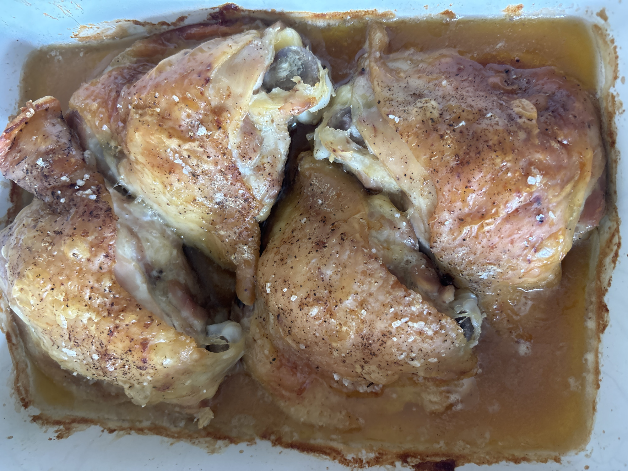 A top-down, close-up shot shows several pieces of roasted chicken, likely thighs and drumsticks, arranged in a white baking dish. The chicken pieces have a golden-brown, slightly crispy skin and are seasoned with visible salt crystals and black pepper. They are partially submerged in a clear, light-yellow liquid, presumably chicken juices or drippings, which fills the bottom of the dish. The lighting is bright and appears to be natural, casting some soft shadows. There are no people visible in the image. The setting is domestic, likely a kitchen or dining area. The image does not contain any information that would allow for the determination of its location in Bila Tserkva, Ukraine.