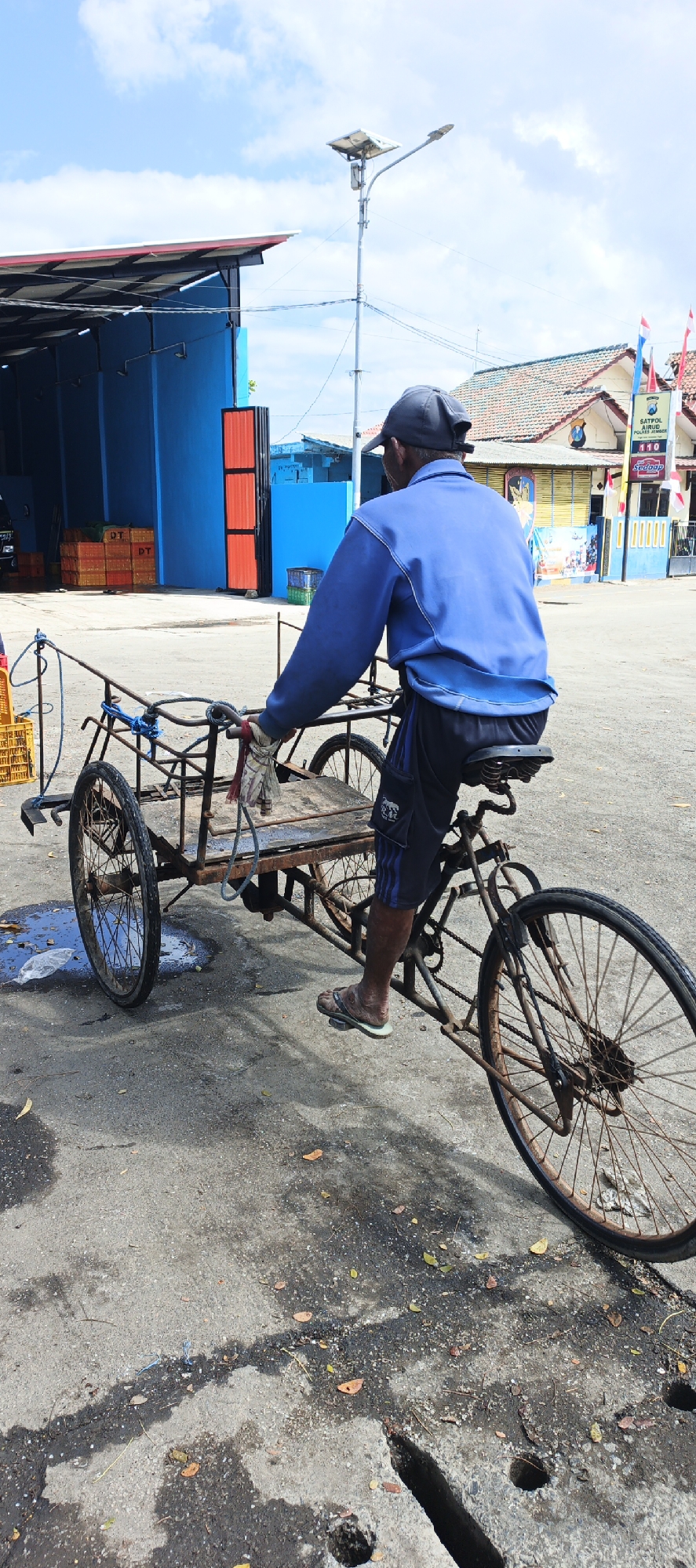 An elderly Indonesian man wearing a blue jacket, black shorts, a dark cap, and sandals is riding a three-wheeled cargo bicycle. The bicycle has a wooden platform in the front and two large rear wheels. He is facing away from the camera. The scene appears to be an outdoor market or port area in Puger, Indonesia, with a bright blue building with red accents and a large awning to the left. Yellow crates are stacked near the building. In the background, there are buildings with tiled roofs and flags. The ground is asphalt with patches of water and fallen leaves. A lamppost with solar panels stands in the background.