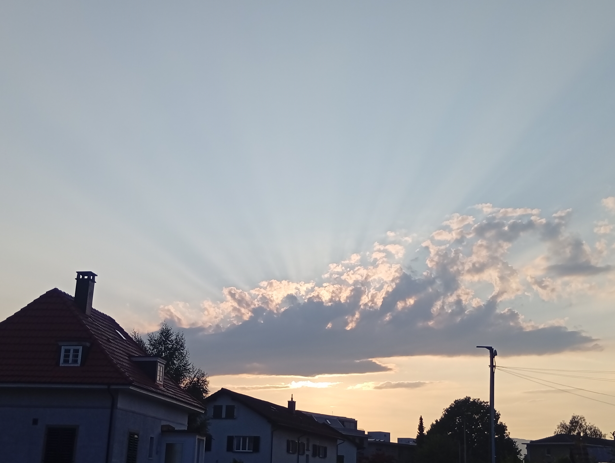 The image, captured in Langenthal, Switzerland, depicts a twilight sky with dramatic crepuscular rays emanating from behind a line of clouds. The sun, low on the horizon, casts a warm, golden glow that tinges the sky and clouds. In the foreground, the roofs and upper portions of residential buildings are visible. One house on the left has a dark red tiled roof with a prominent chimney and dormer windows. Another house further to the right has a darker roofline. Trees are interspersed between the buildings. A lamppost with wires is visible in the middle right of the frame. The overall scene suggests the end of a day, with natural light playing a significant role in the visual composition.
