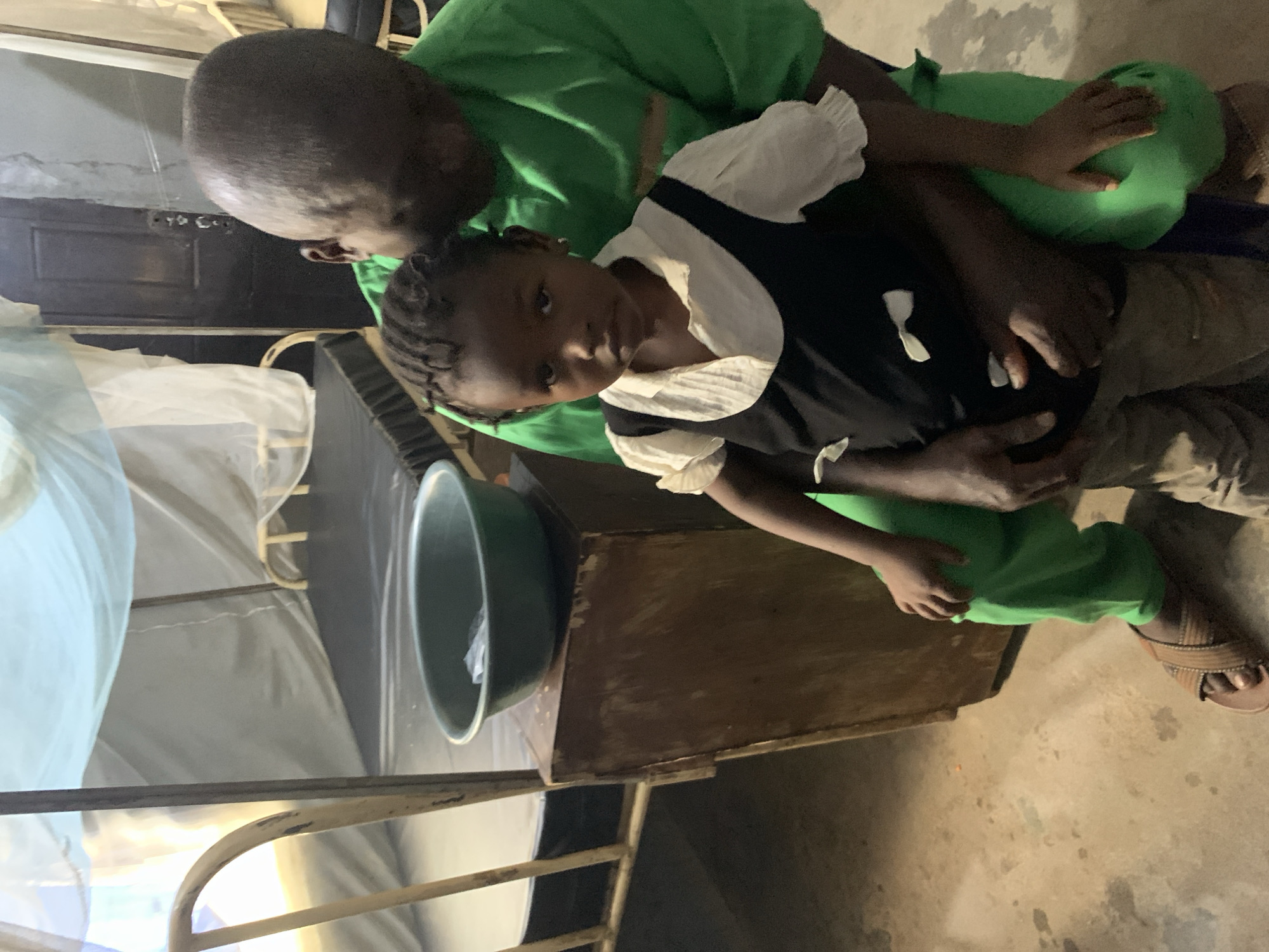 The scene is in what appears to be a room, likely in Akwanga, Nigeria. An adult wearing a green shirt is holding a young child, who is wearing a black and white dress. A metal bed frame is visible to the left, partially covered with a white, tent-like netting. A dark-colored basin or container is sitting on a wooden surface near the bed. The floor is light brown and appears to be concrete or a similar material. The room is dimly lit.