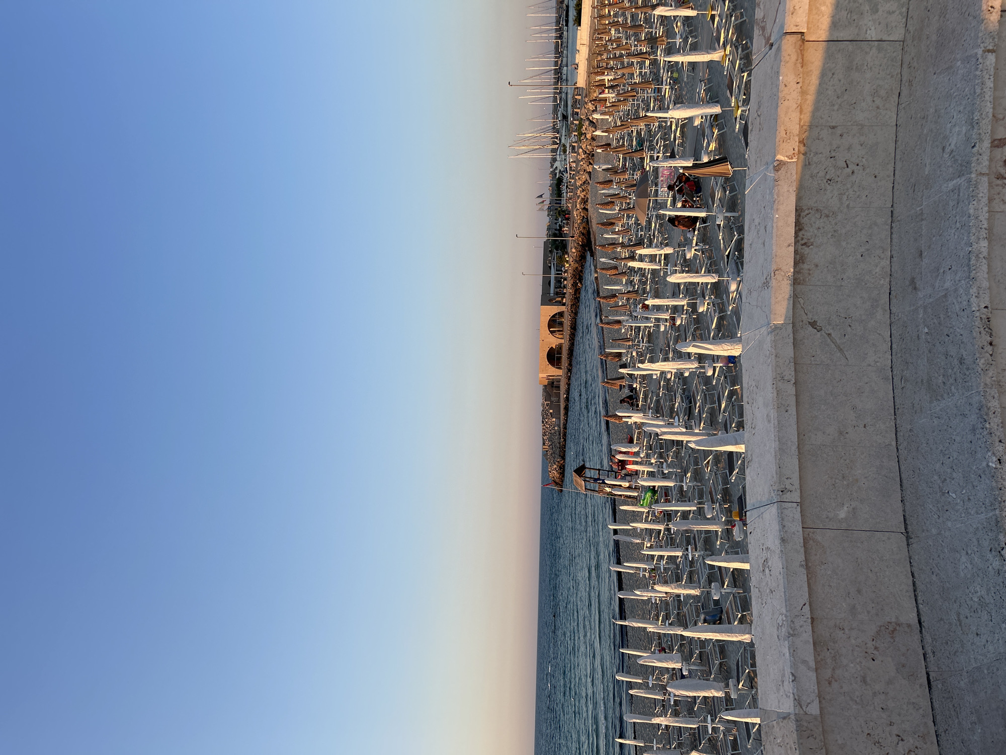 The image captures a sunset on a beach in San Foca, Italy. The sky is a clear gradient of light blue to pale orange. The sea, to the left, is calm with gentle waves. In the foreground, a stone wall with a textured surface lines the elevated viewpoint.
The beach is densely populated with rows of white and beige lounge chairs and closed umbrellas, arranged in a grid-like pattern. A lifeguard stand with a red flag is visible in the middle of the beach, and a few people are seated on the lounge chairs or walking in the distance.
Further back, a man-made jetty extends into the sea, topped with a modern building featuring arched windows. Beyond the jetty, a marina is visible with numerous sailboats docked. The overall scene conveys a peaceful, end-of-day atmosphere on the Adriatic coast.