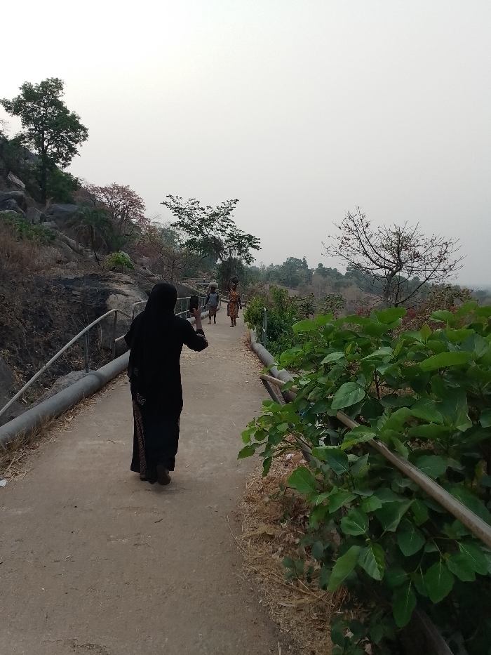 A woman in a black robe stands on a concrete path, holding a phone as if taking a photo. She faces away from the viewer, looking towards two women carrying items on their heads in the distance. The path is bordered by railings and greenery on the right, and a rocky slope with trees on the left. The sky is overcast, suggesting either early morning or late evening.