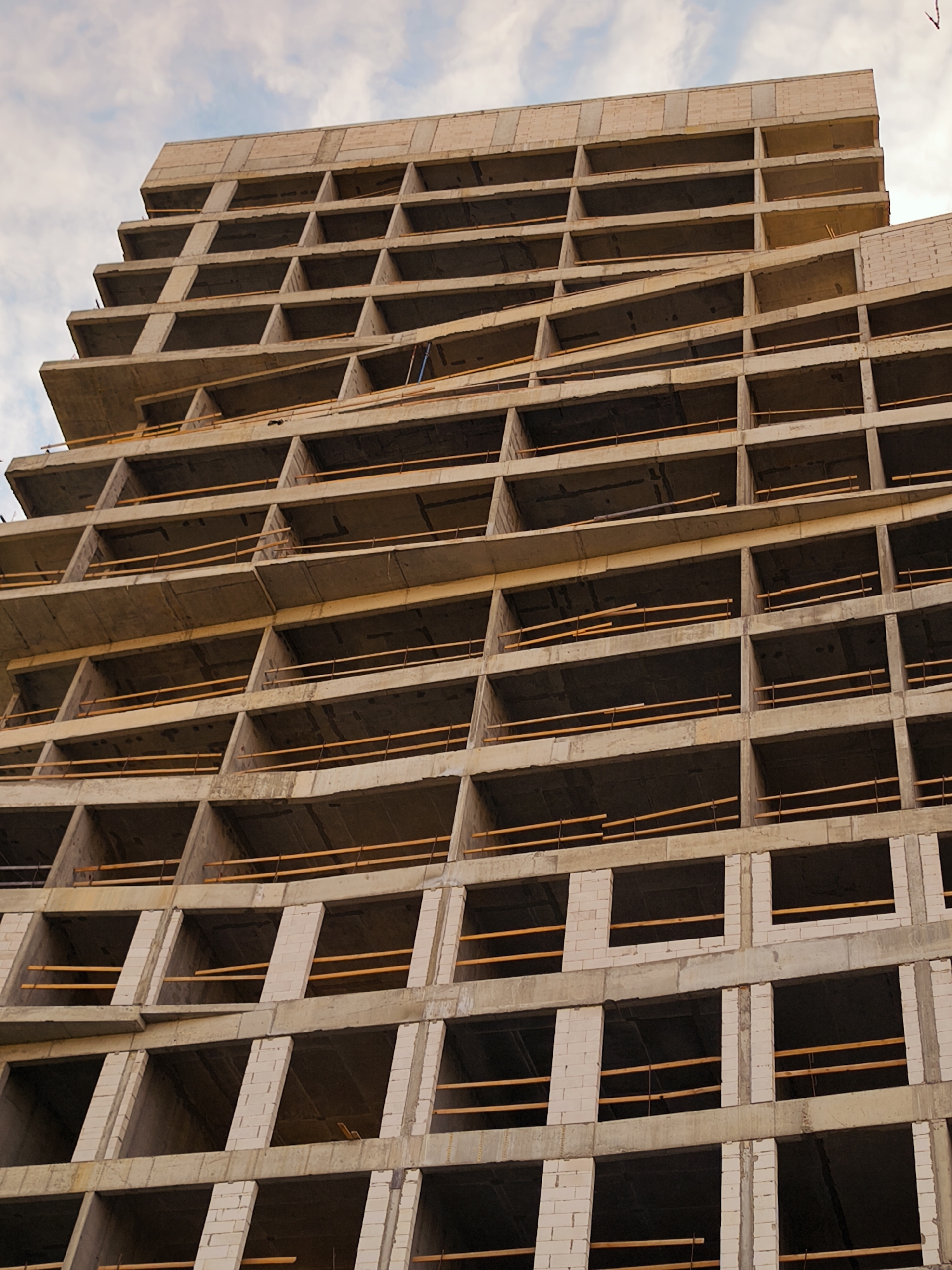 The image shows a tall, unfinished building with a concrete frame and exposed floors. The building has a grid-like structure with many open rectangular openings that appear to be window or balcony spaces. Wooden planks are visible across some of these openings, possibly acting as temporary barriers or supports. The concrete surfaces are rough and show signs of construction. The sky above is partly cloudy. The building's design suggests a modern architectural style, likely a residential or office complex under construction. No people are visible in the image. The context of Moscow, Russia, suggests this is likely a new development in a rapidly urbanizing area.