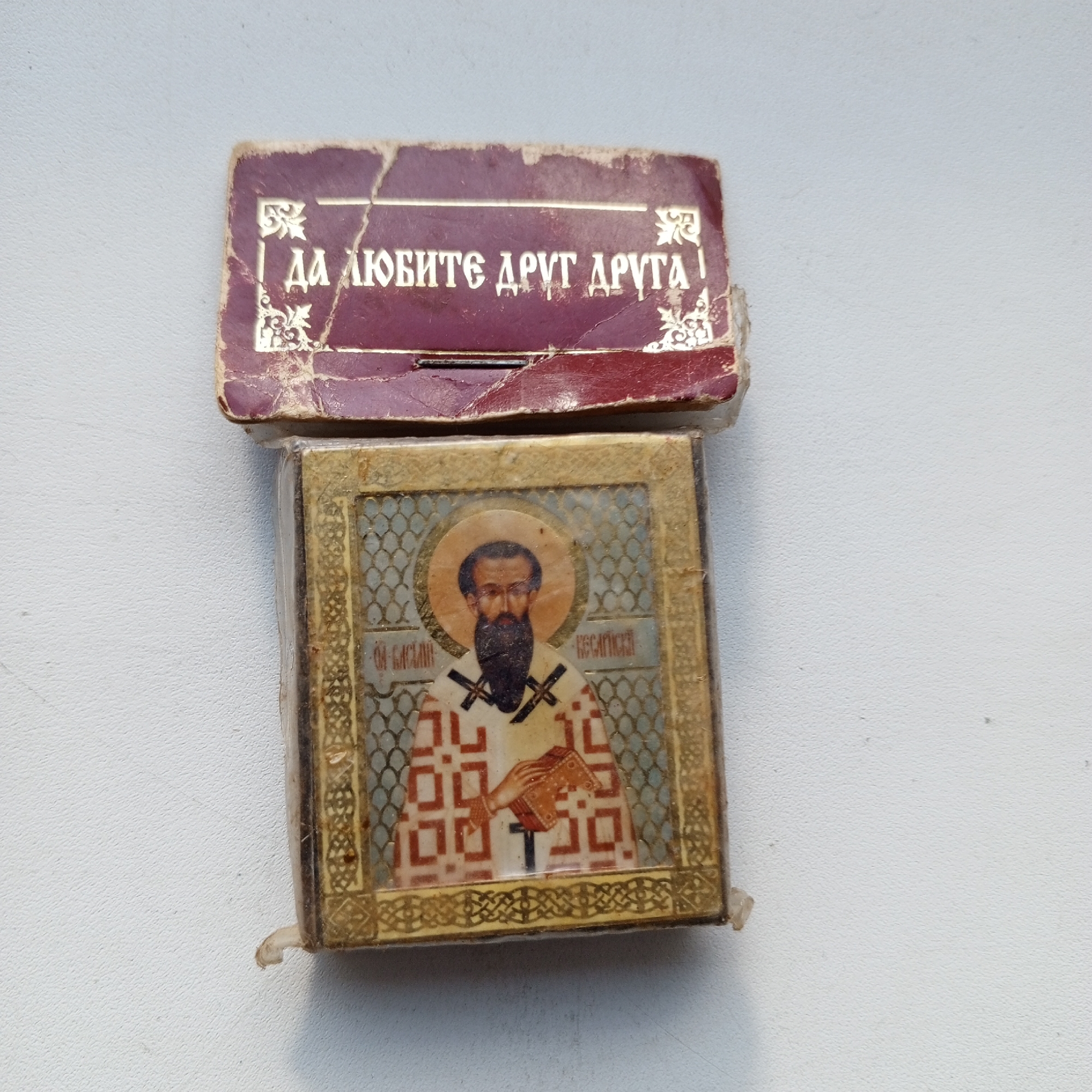 This is a close-up, top-down shot of an open box containing a small, rectangular Orthodox icon. The box is made of dark red material with gold lettering that reads "ДА ЛЮБИТЕ ДРУГ ДРУГА", which translates to "Love one another". The box is worn and torn, with visible damage to its edges. The icon inside depicts a male saint with a beard, wearing religious vestments and holding a book. The icon is encased in plastic packaging, which is slightly creased. The background is a plain, textured white surface. The items are on a white, textured surface. The image does not contain any identifiable information about the location in Sayanogorsk, Russia.
