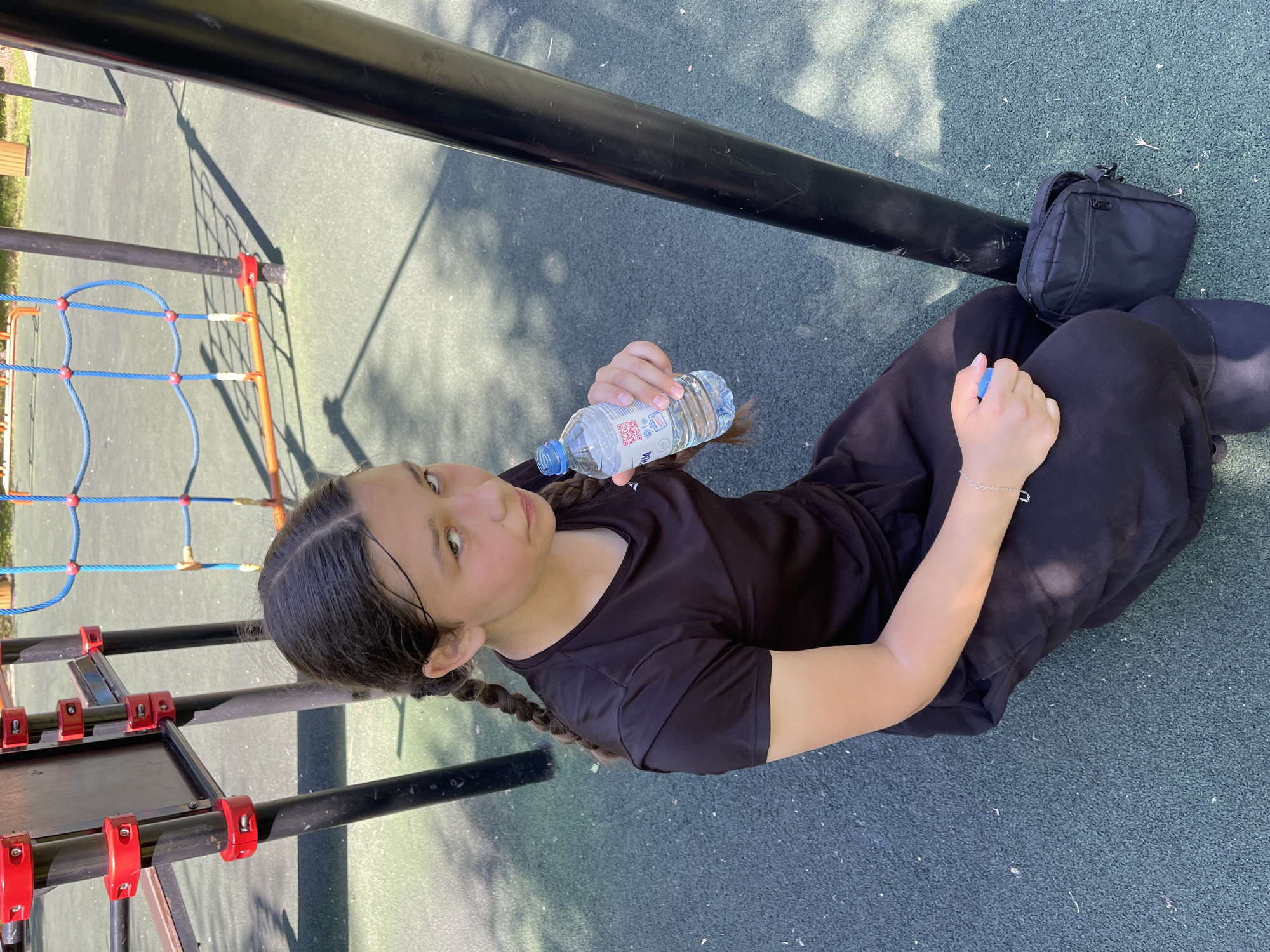 A young girl with dark hair in braids sits on a dark green rubberized playground surface, holding a plastic bottle of water. She is wearing a black t-shirt and black pants. To her right is a small black bag with a zipper. In the background, playground equipment with red and blue accents is visible. The overall environment suggests an outdoor recreational area.