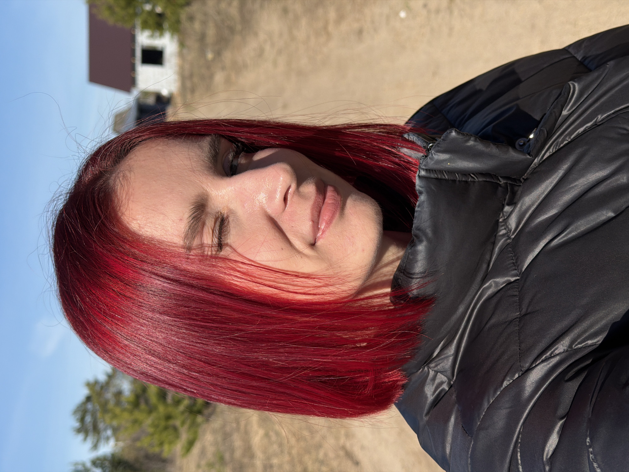 A woman with vibrant red hair is posing outdoors. She is wearing a black puffer jacket. The background features a beige-colored ground, trees, and a building. The image was taken during daylight hours in Chaikovsky, Russia.