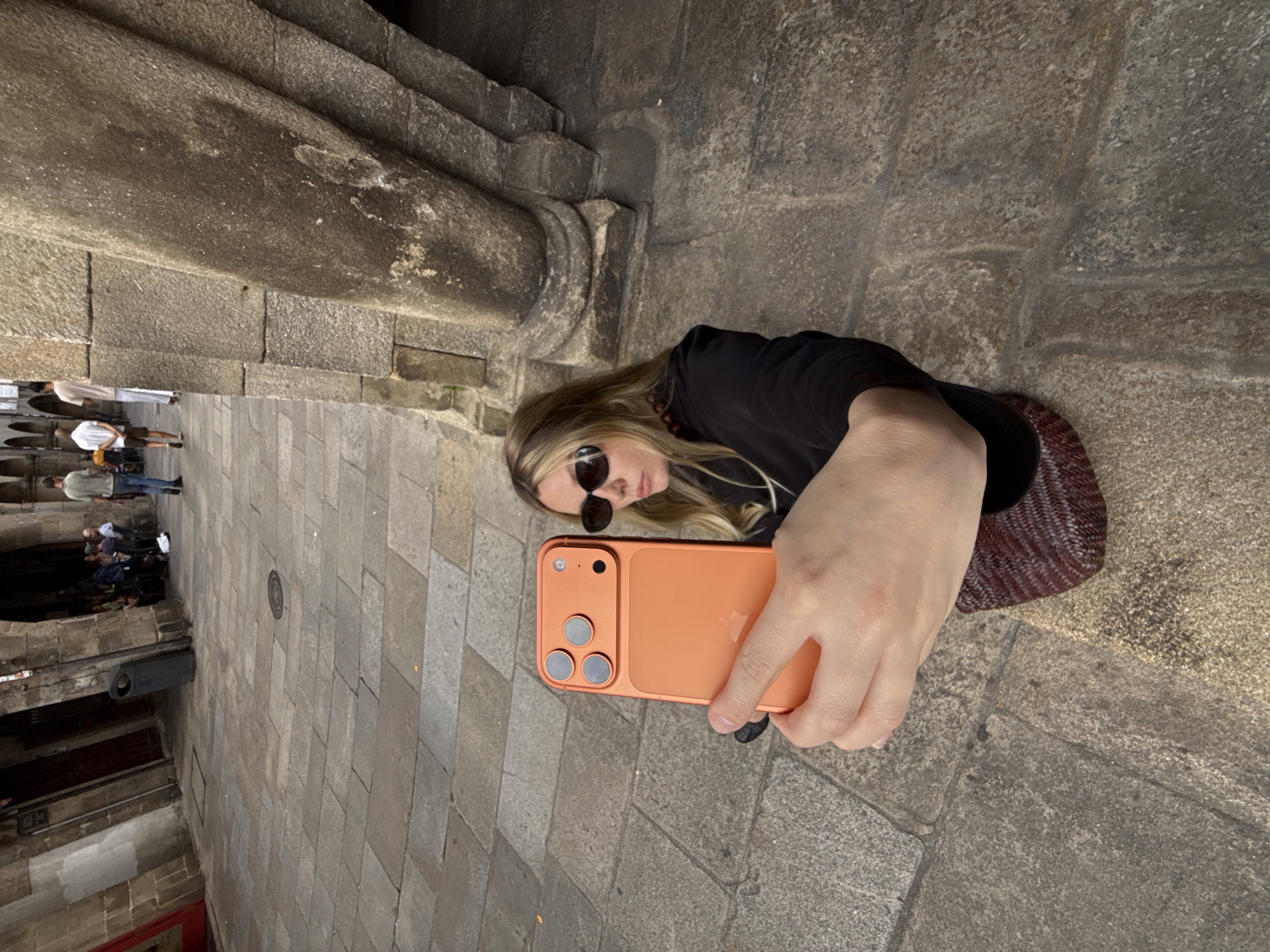 A woman is taking a selfie with an orange smartphone in an outdoor courtyard in Santiago de Compostela, Spain. The courtyard is paved with large, grey stone slabs, and there are stone arches and columns in the background. Several people are walking in the distance, and some are seated at tables in an outdoor cafe. The woman is wearing sunglasses and a black top. The smartphone has a triple-lens camera system.