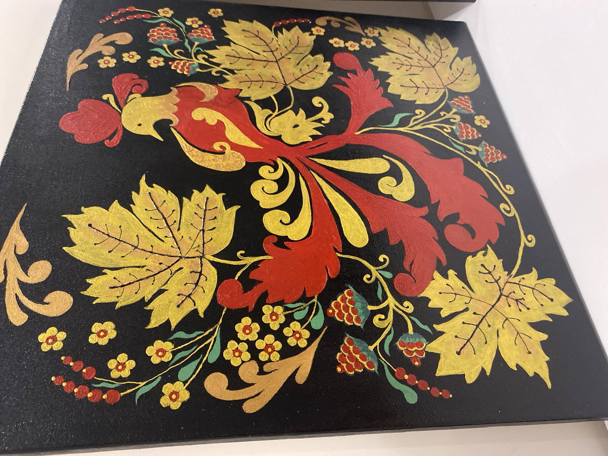 The image shows a close-up, angled view of a black canvas painting with vibrant, stylized folk art. The painting features a central motif of a golden and red bird with a red crest and elaborate tail feathers. Surrounding the bird are large, golden leaves with black veins, small red berries on green stems, and clusters of small yellow and red flowers. The overall style is reminiscent of Russian Khokhloma painting, characterized by its bold colors and decorative patterns against a dark background. The painting is displayed on a white surface, likely a shelf or table, with a plain white background. The lighting is bright, highlighting the texture of the canvas and the glossiness of the paint. The exact location in the city of Unknown, Unknown is not specified in the image.