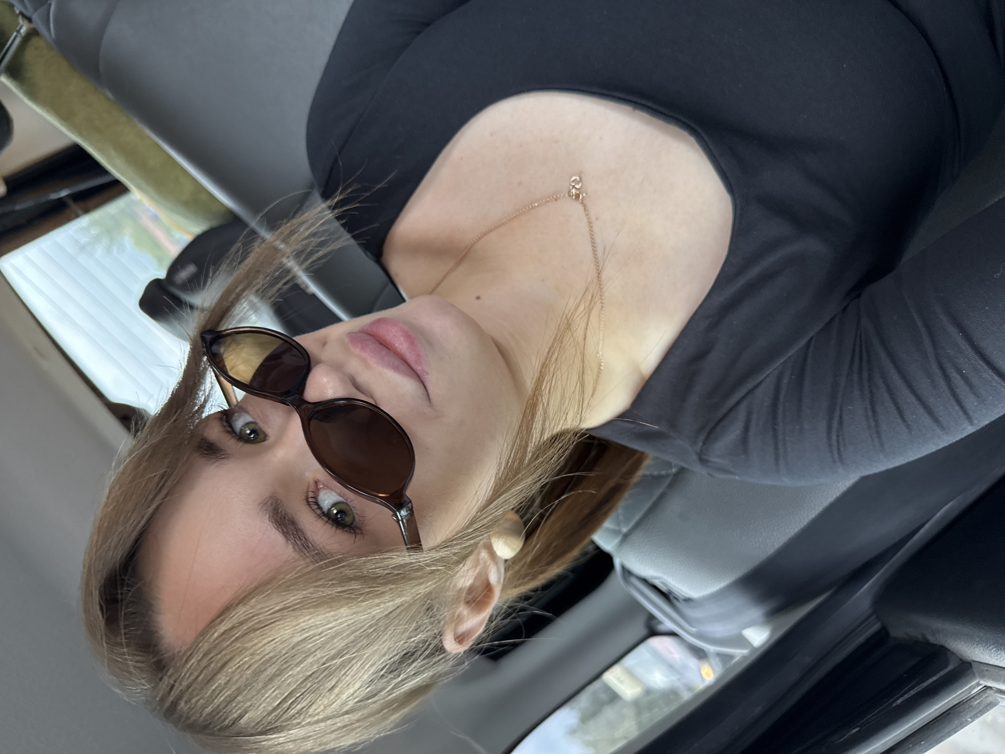 In the city of Chaikovsky, Russia, a blonde woman with light brown hair pulled into two pigtails and bangs is pictured inside a vehicle. She is wearing dark sunglasses, a black top, and a delicate gold necklace with a pendant. The interior of the vehicle is visible, with grey seats and a glimpse of the outdoors through the windows. The image is a close-up, focusing on the woman's face and upper body.