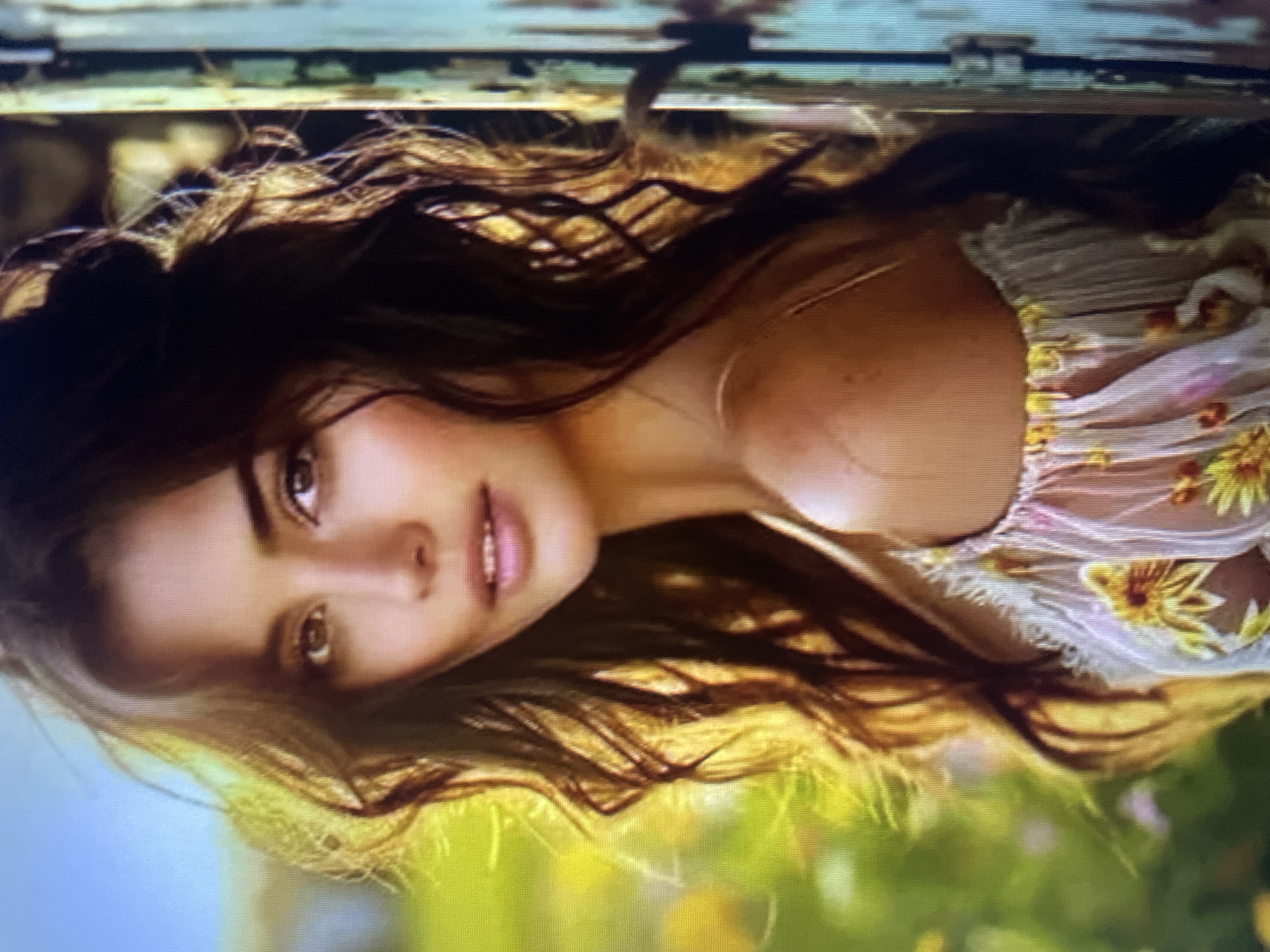 A medium shot captures a young woman with long, dark, wavy hair against a blurred background of green foliage and yellow flowers, illuminated by warm, golden sunlight. She is positioned to the left of a rustic, pale blue wooden structure with visible vertical lines and a dark hinge on the right side of the frame. The woman is looking towards the viewer with a gentle expression, her head tilted slightly. She is wearing a light-colored, off-the-shoulder garment with a ruffled neckline adorned with floral patterns of yellow and hints of pink. The image does not provide any information about the city of Kołobrzeg, Poland.