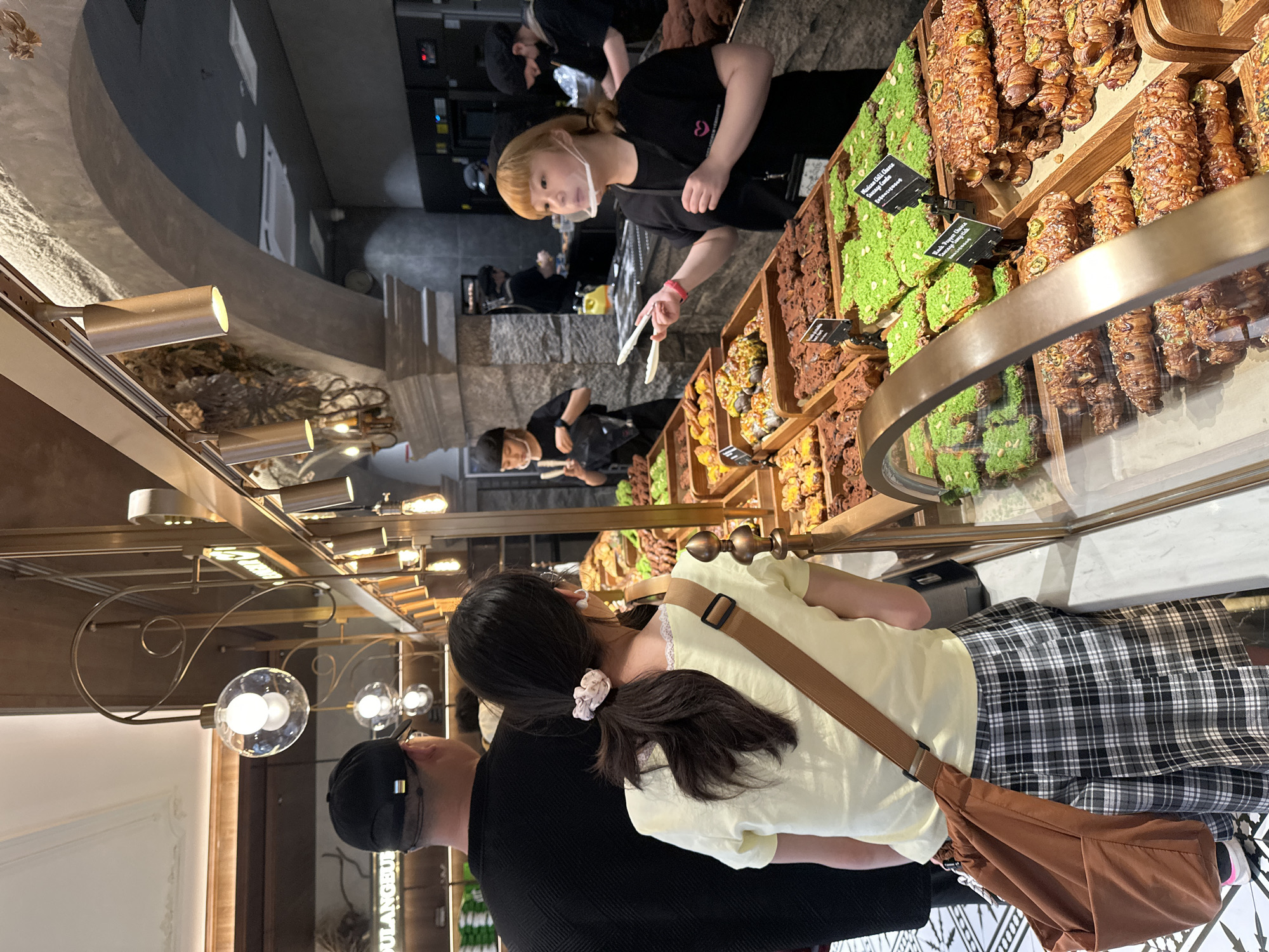 In Chongqing High-tech Zone, China, a bakery scene unfolds. An employee, a young woman with light blonde hair wearing a black shirt and a face mask, uses tongs to handle baked goods. Patrons, including a person wearing a black hat and shirt, along with a woman in a yellow shirt and plaid skirt with a brown shoulder bag, are visible, potentially customers. The environment includes a glass display case filled with various pastries, along with ornate light fixtures hanging from the ceiling.